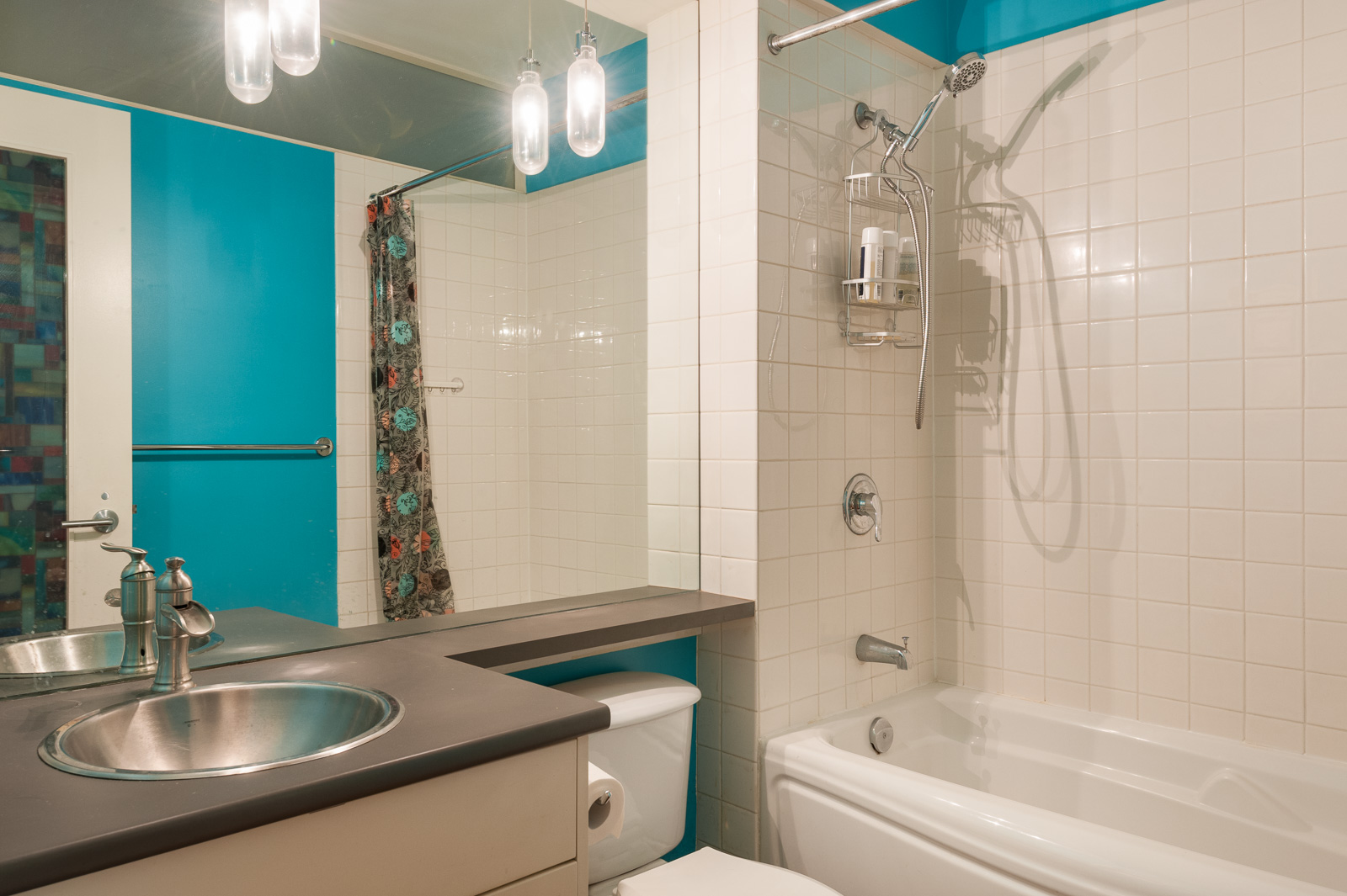 Modern bathroom with turquoise walls, white tiles, a bathtub-shower combo with a curtain, a sink with a metal faucet, and a large mirror above the counter.
