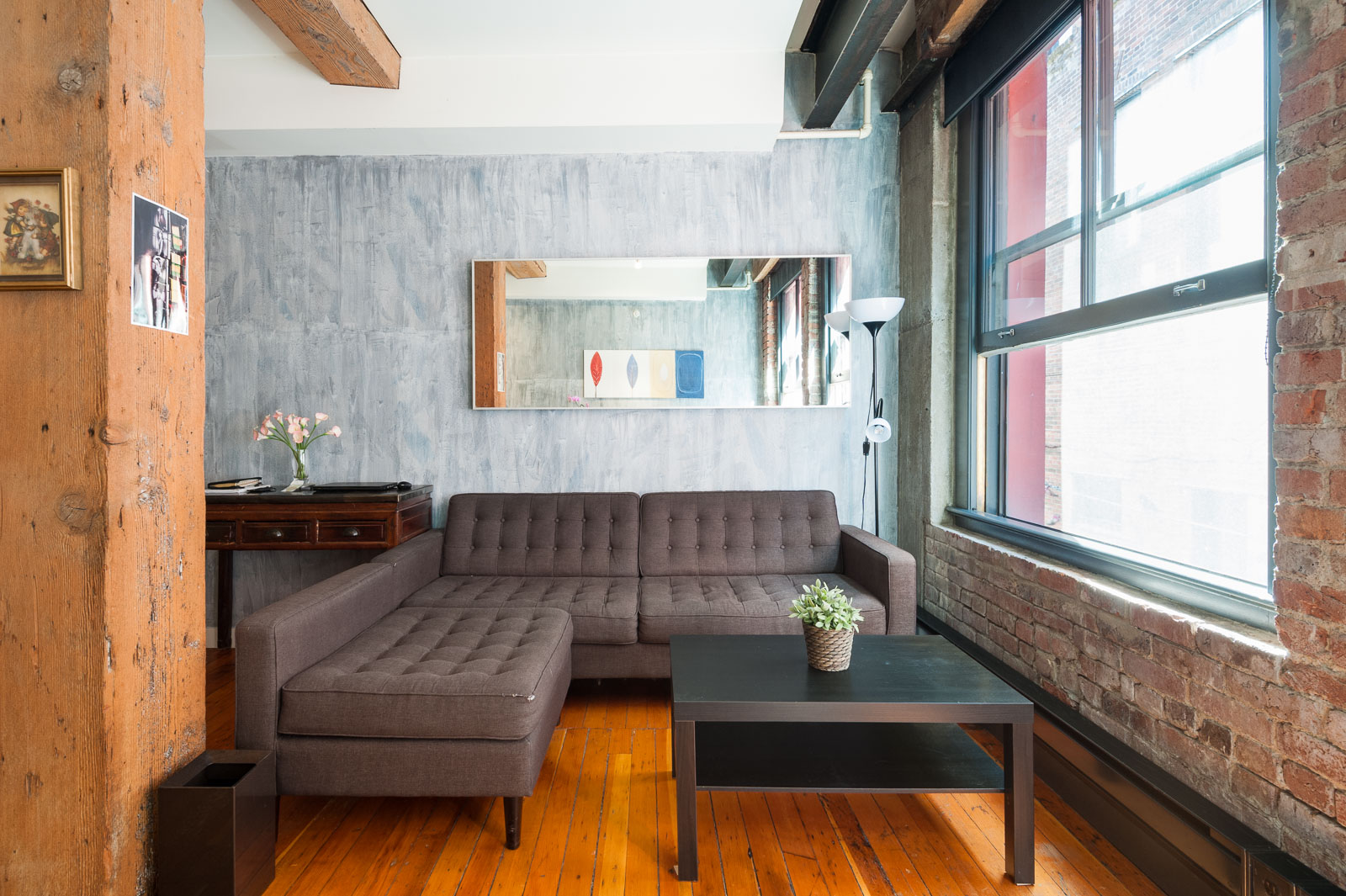 Modern living room with a gray sectional sofa, black coffee table, and a large window; exposed brick walls and wood beams add industrial style.