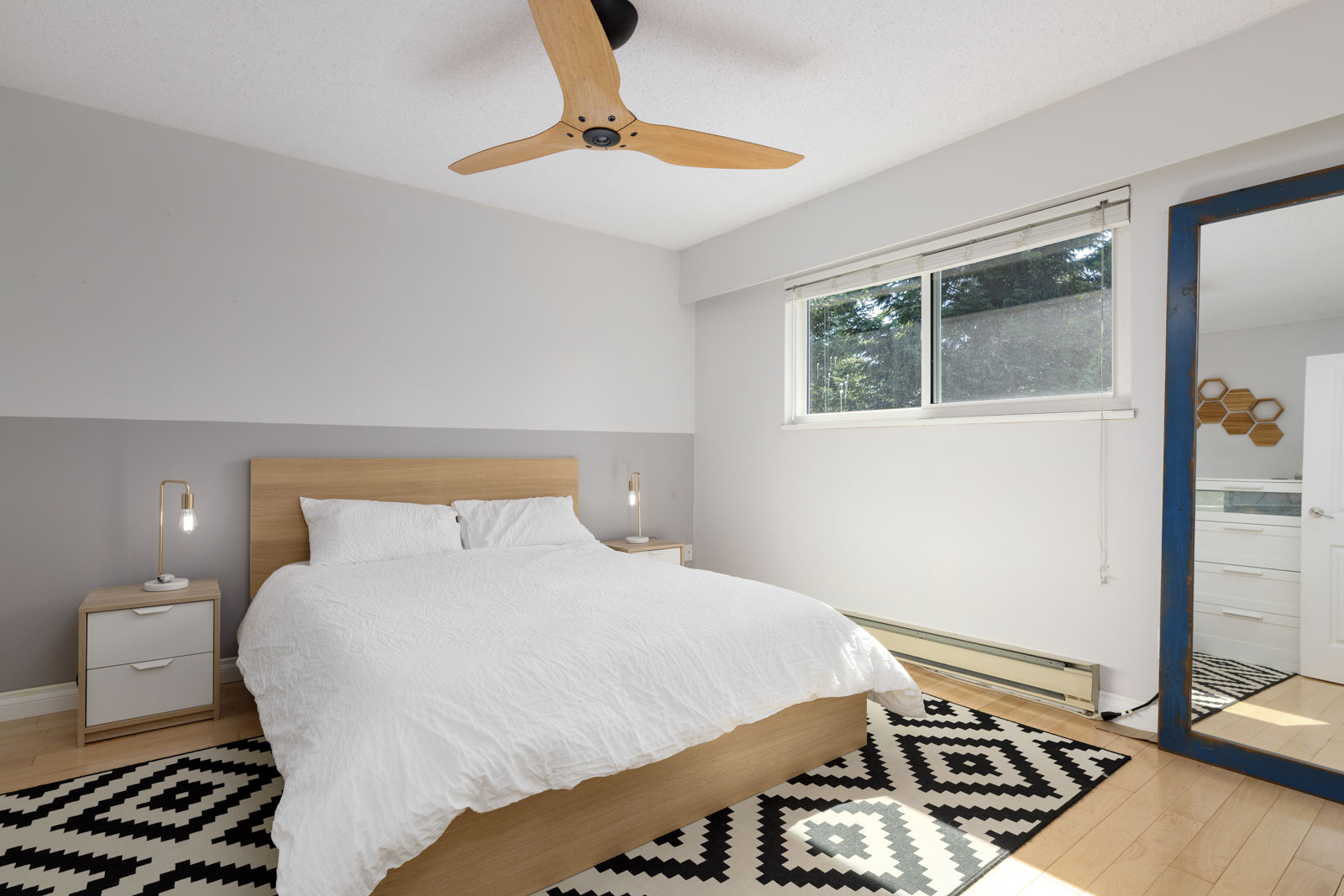 Modern bedroom with a wooden bed, white bedding, bedside tables, ceiling fan, geometric rug, large window, and a mirror reflecting an adjacent room.