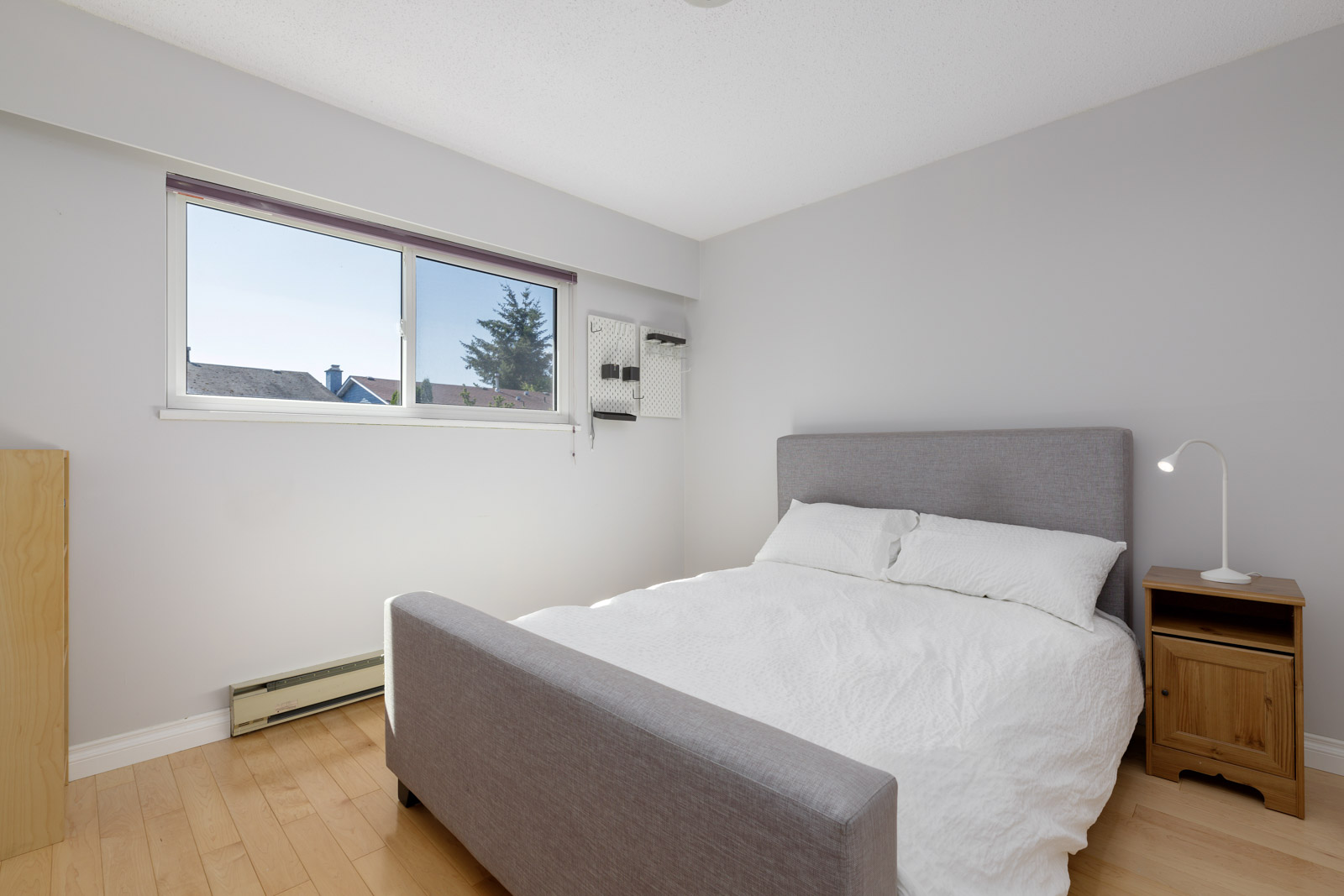 A small bedroom with a gray upholstered bed, white bedding, a wooden nightstand with a lamp, light wood floors, and a window letting in daylight.