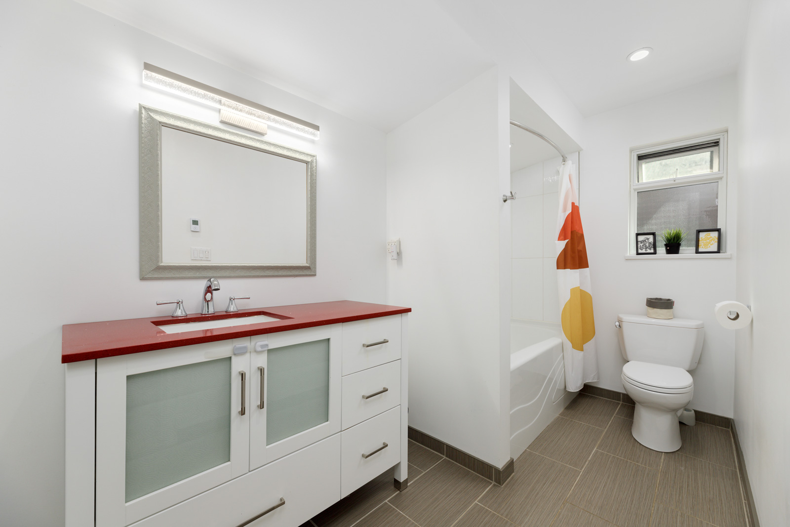 Modern bathroom with a red countertop vanity, white cabinets, a wall mirror, toilet, bathtub with shower curtain, and small window above the toilet.