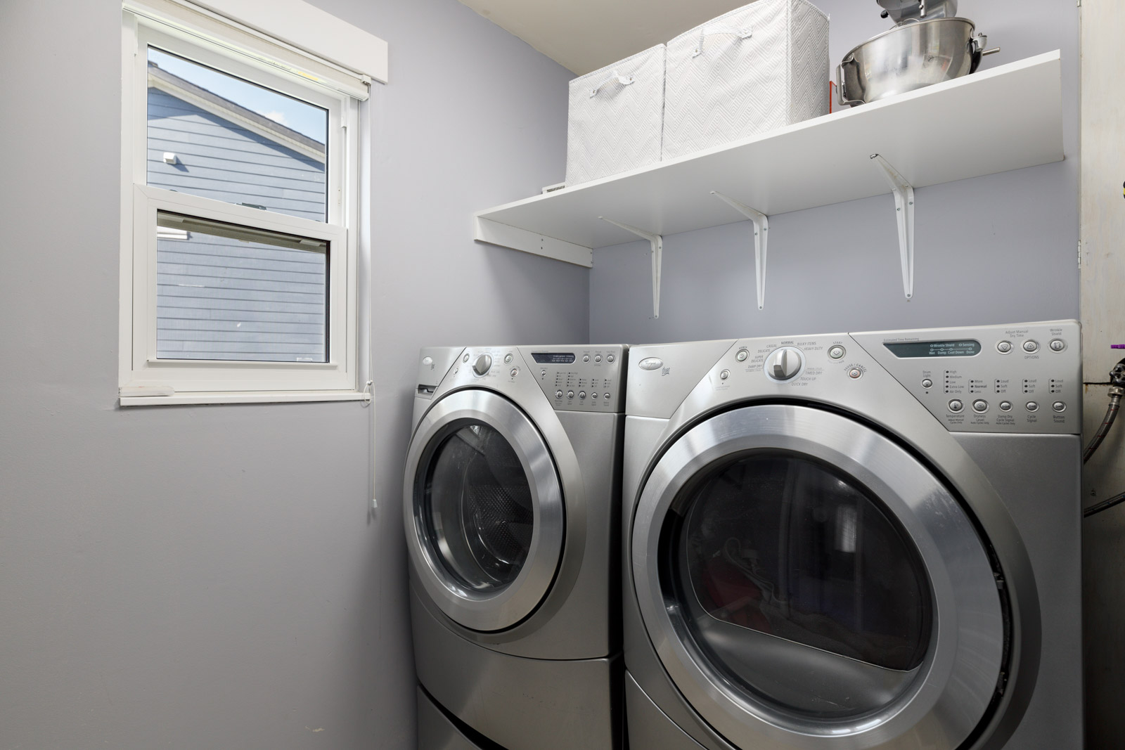 Modern laundry room with a front-loading washer and dryer, open shelf above holding two boxes and a stand mixer, and a window to the left.