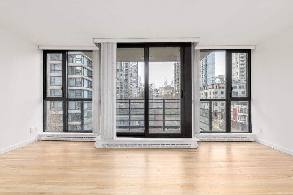 Empty modern room with light wood floors, large windows, and sliding glass doors overlooking tall city buildings.
