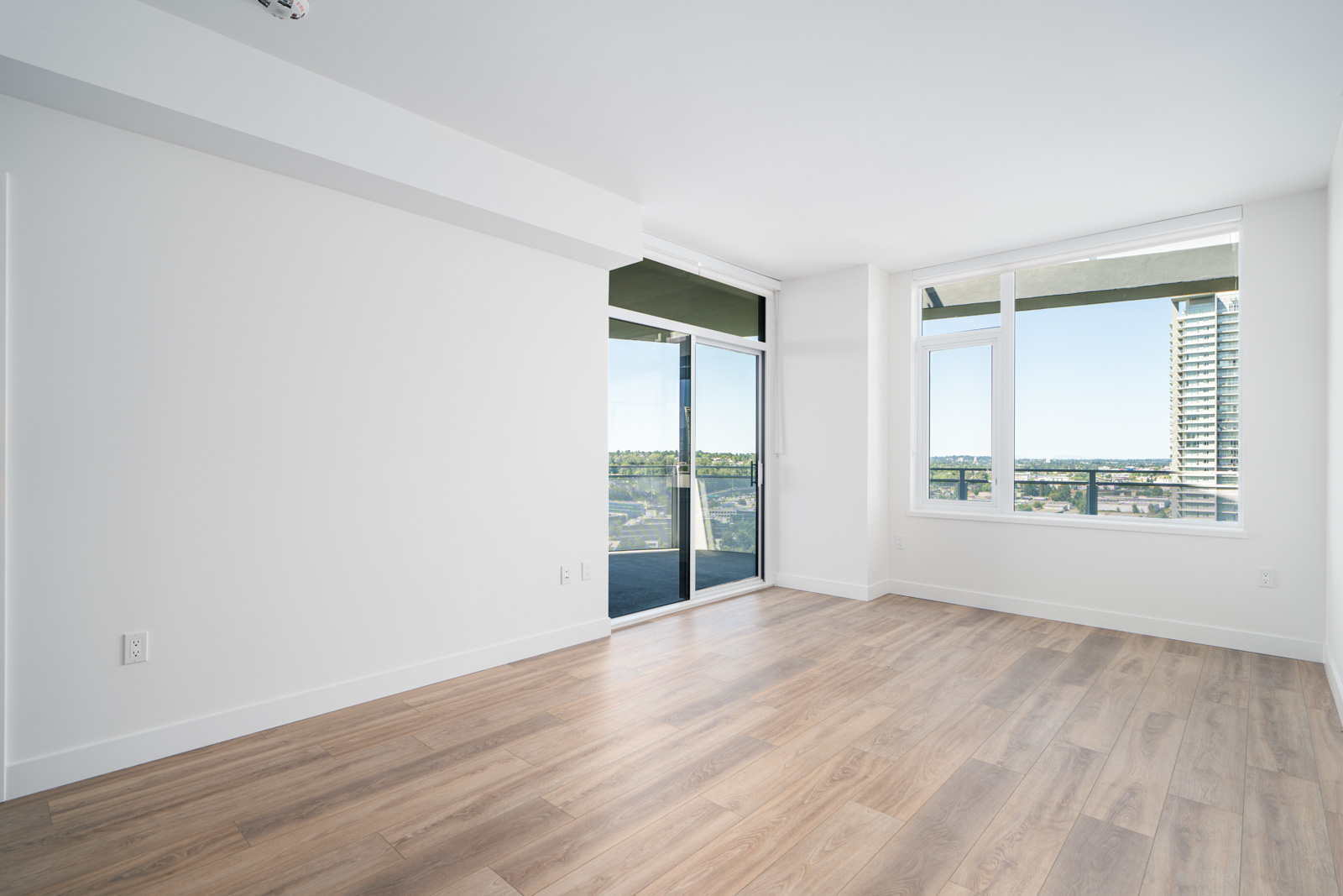 Empty modern room with light wood flooring, white walls, large windows, and a glass door leading to a balcony with a city view.
