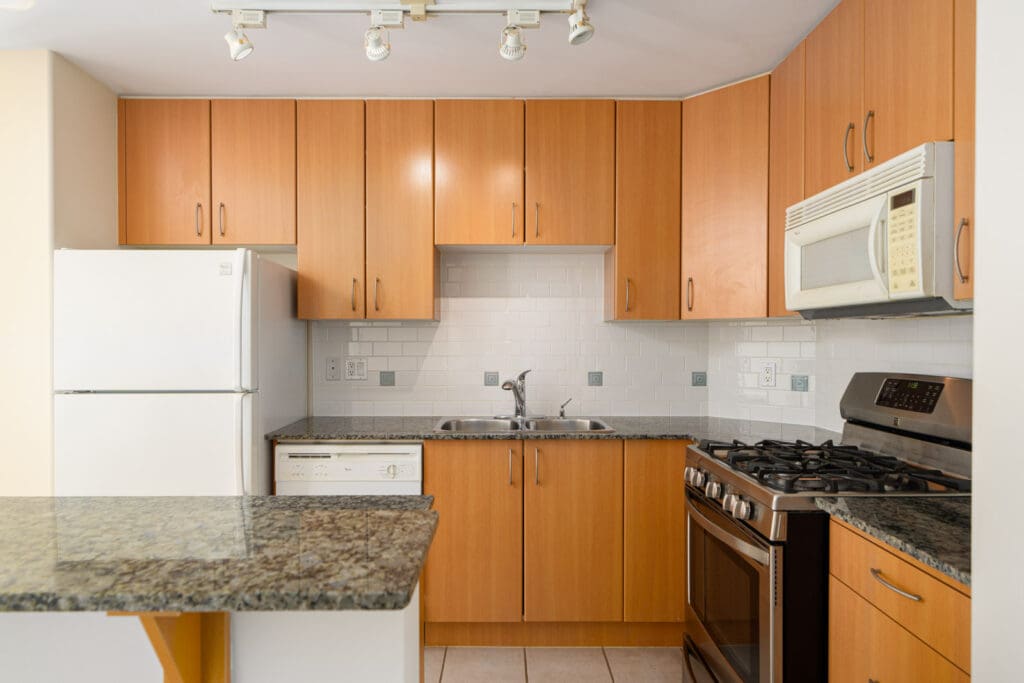 Modern kitchen with wood cabinets, granite countertops, stainless steel gas stove, white refrigerator, microwave, and dishwasher, all set against a tiled backsplash.