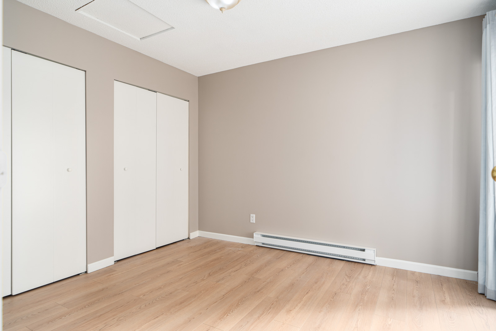 A room with light beige walls, light wood flooring, two white closet doors, a baseboard heater, and an overhead light fixture.