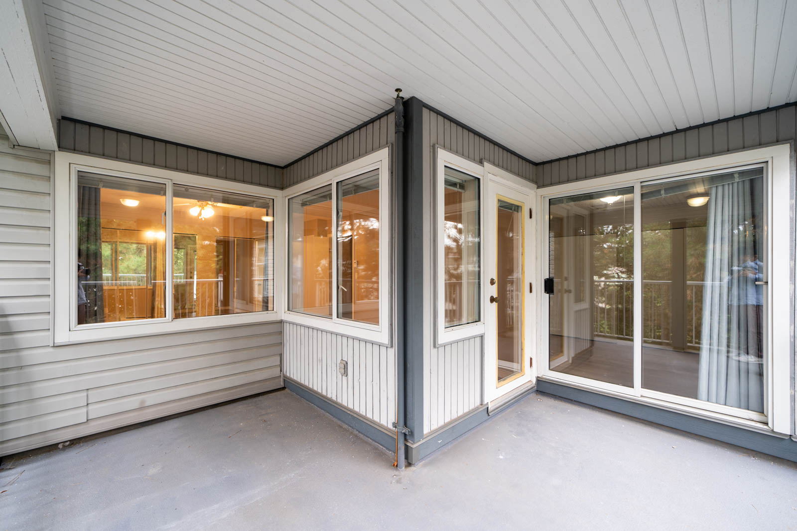 A covered outdoor corner patio with large windows and a sliding glass door. The interior lights are on, and the patio is surrounded by a grey wall with a white ceiling.