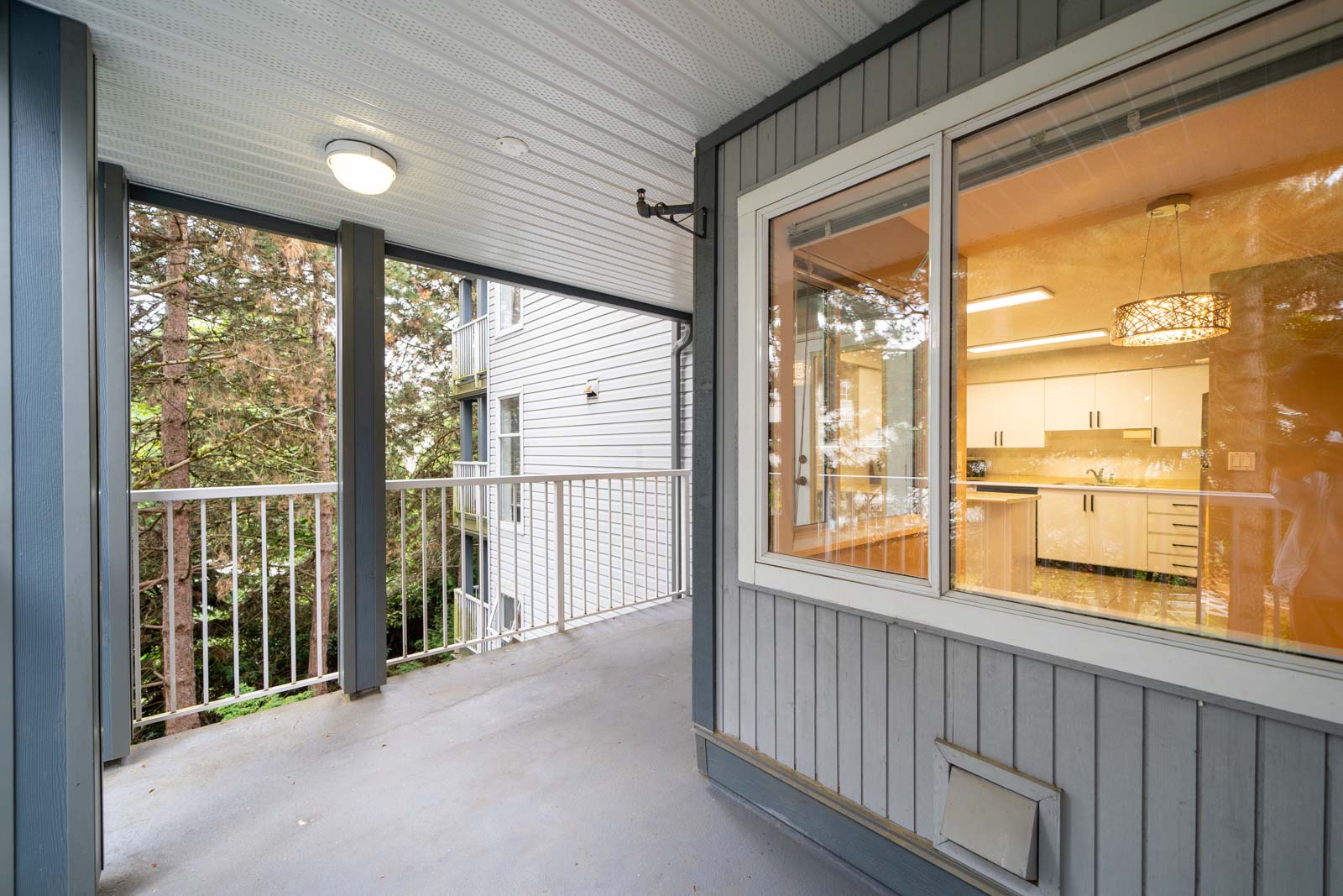 A covered balcony with gray paneling, a white railing, and a view into a kitchen area through a large window. The balcony has a ceiling light and overlooks a wooded area.