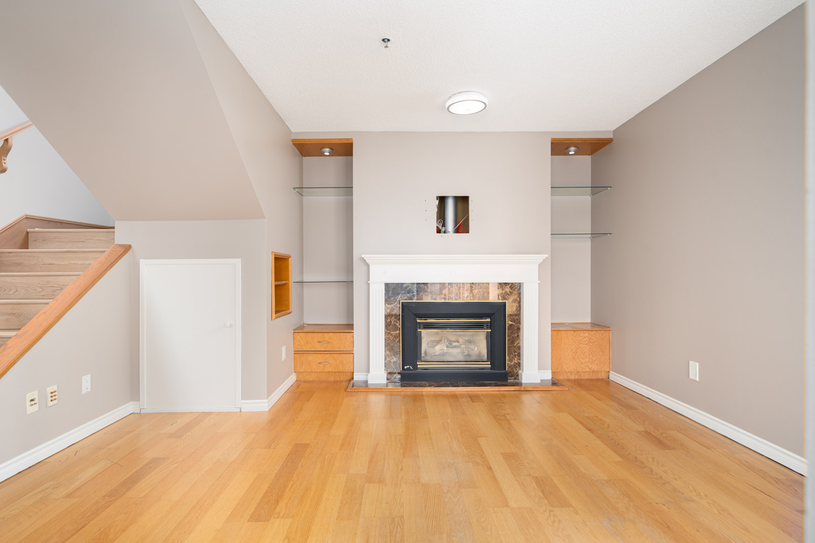 A vacant living room with light wood flooring, a central fireplace, recessed wall shelving, and a staircase to the left. Three wall outlets are visible on the left wall.