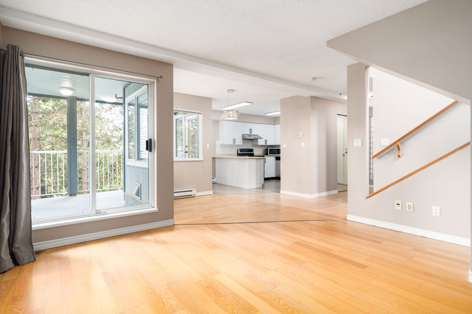 A spacious, modern living area featuring hardwood floors, an attached balcony with sliding glass doors, an open-concept kitchen with white cabinetry, and a staircase leading to the upper floor.