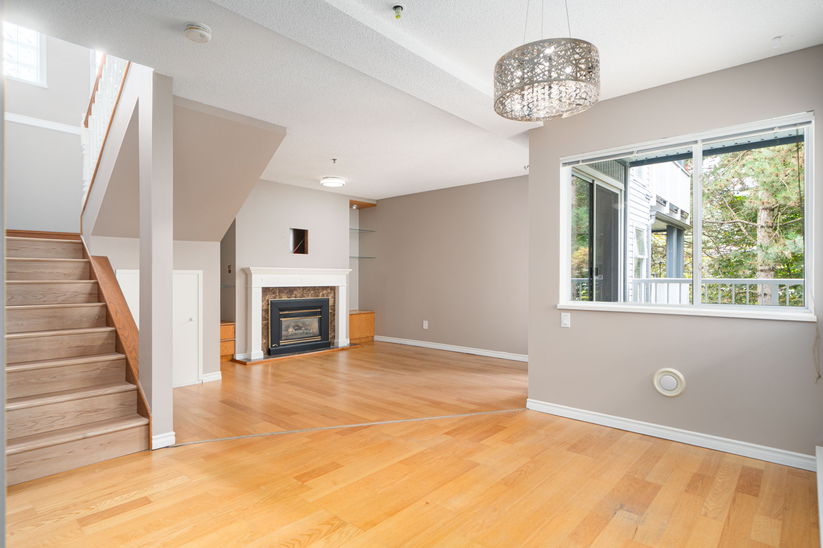 A bright, empty living room with light wood floors, a gas fireplace, a stairway on the left, a window on the right, and a modern chandelier on the ceiling.