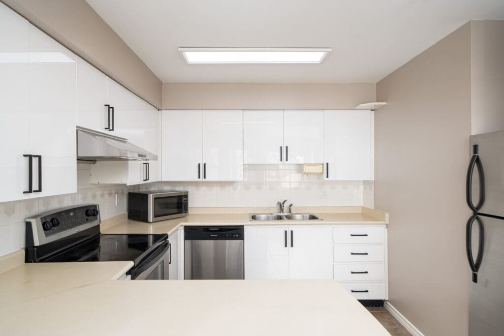 A modern kitchen with white cabinets, black handles, stainless steel appliances including a stove, microwave, dishwasher, and refrigerator, and a neutral beige countertop.