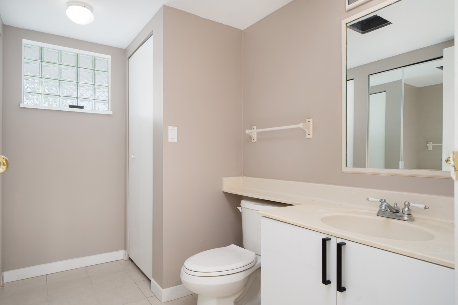 Bathroom featuring a vanity with a sink and mirror, a toilet, a window with frosted glass blocks, and beige walls.