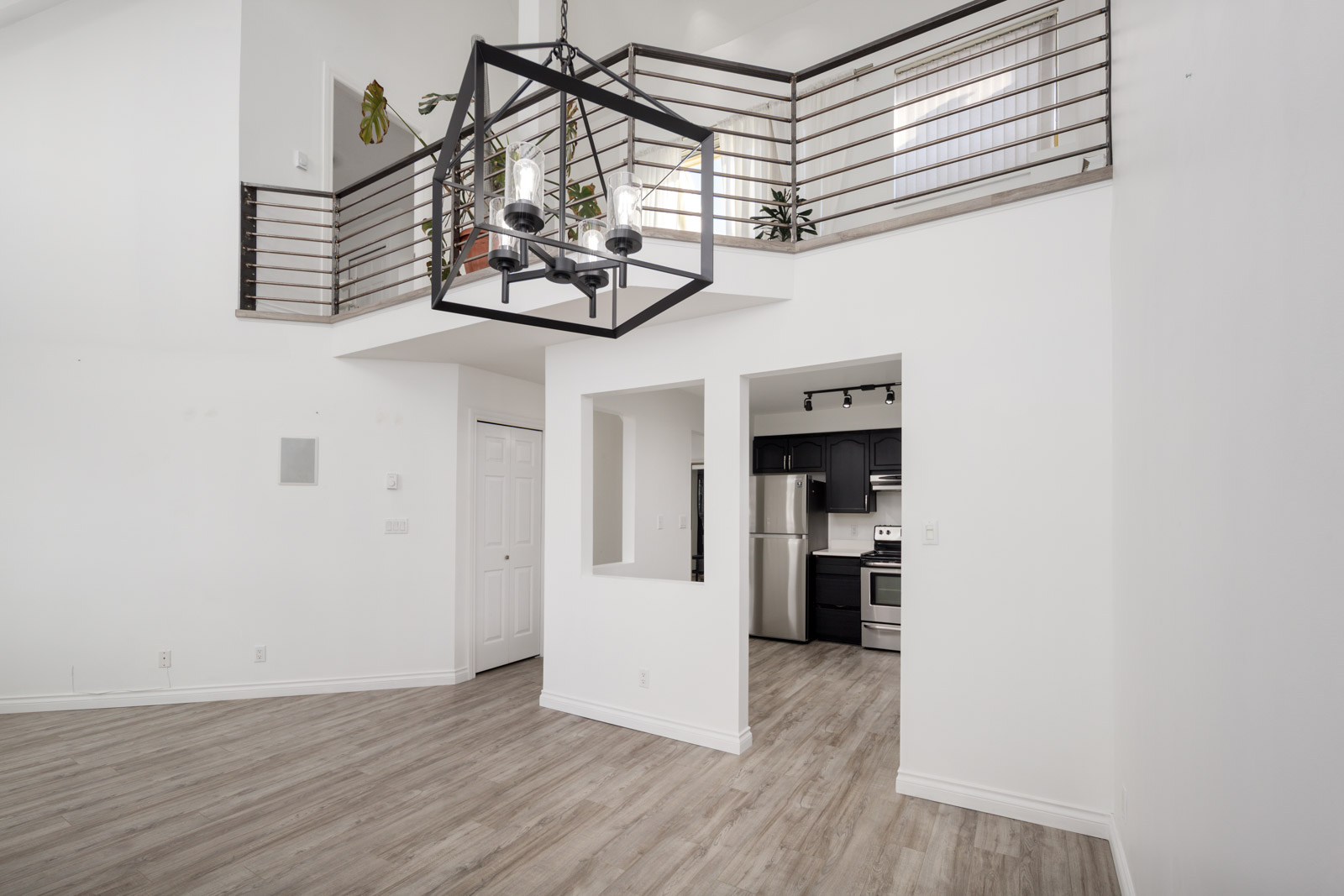 A modern loft interior with white walls, wood flooring, a black metal chandelier, open railing on an upper level, and a kitchen with stainless steel appliances.