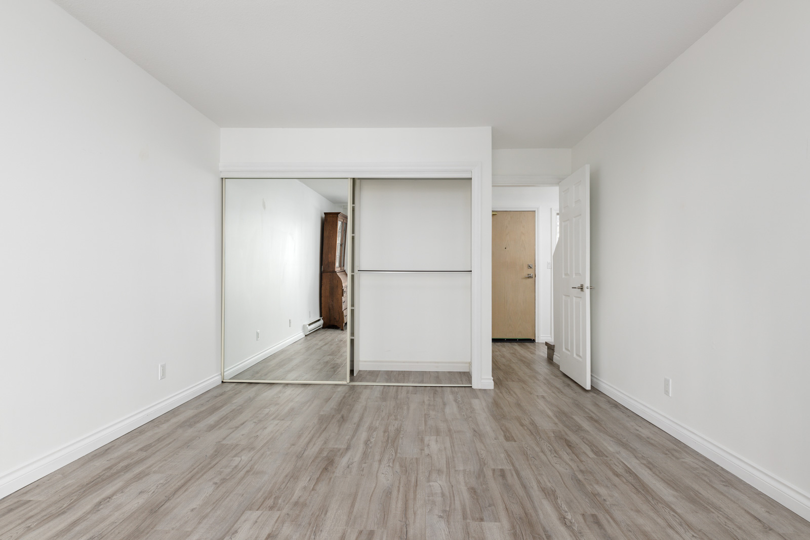 Empty room with light-colored wood flooring, white walls, a large closet with sliding mirror doors, and a doorway leading to an adjacent space.