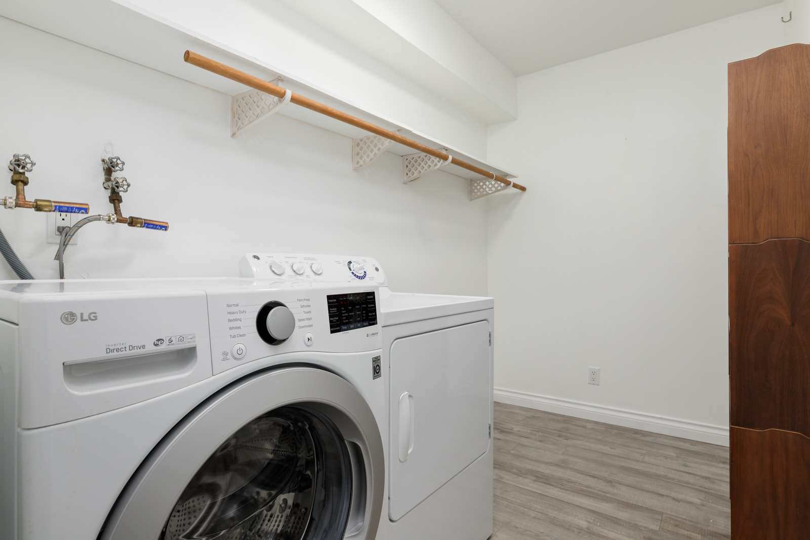 A laundry room with an LG front-load washer and a dryer, light wood flooring, white walls, and a wooden shelf with white baskets above the machines.