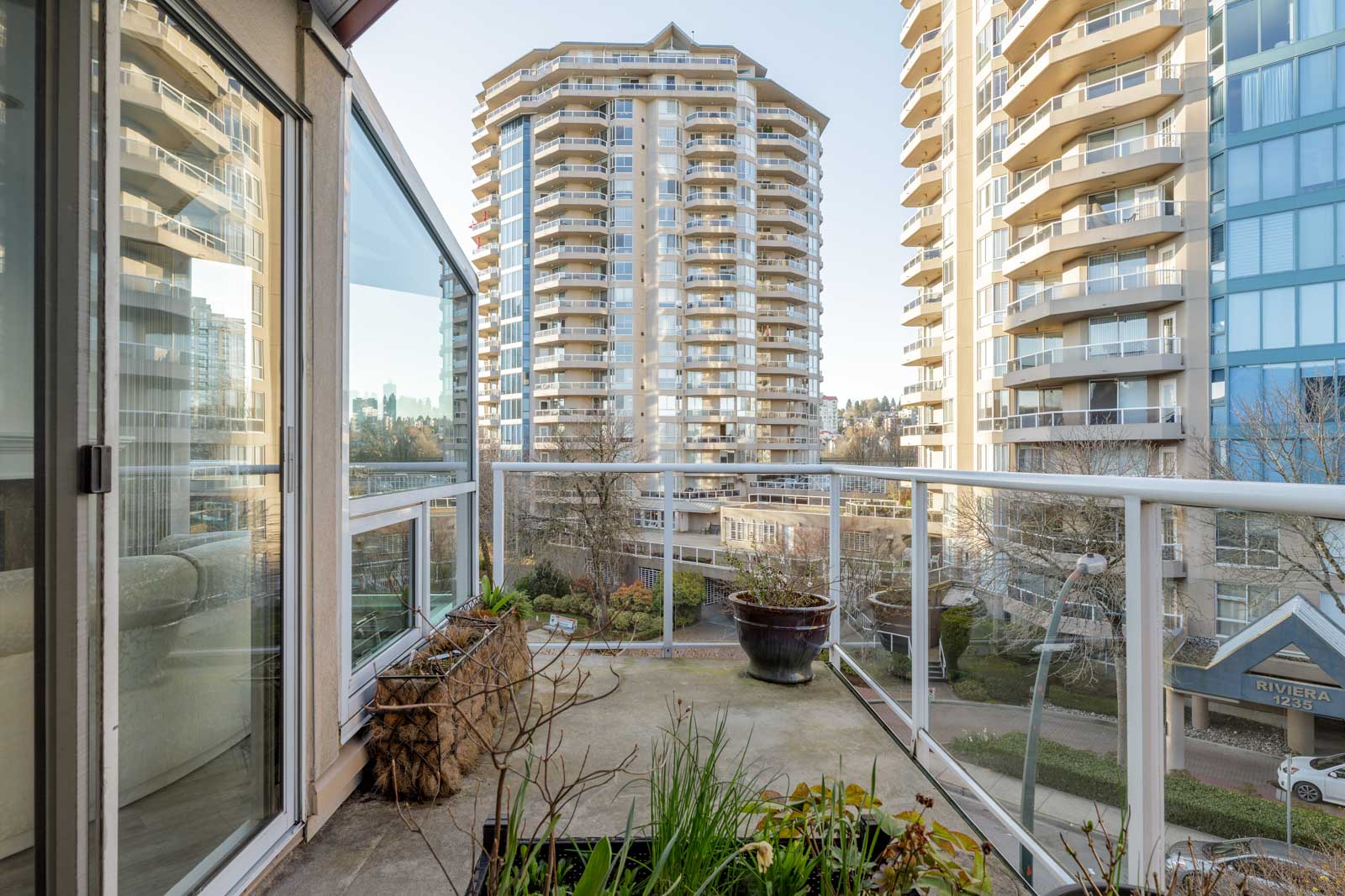 View from a balcony showing nearby high-rise apartment buildings, potted plants, and glass railings on a clear day.