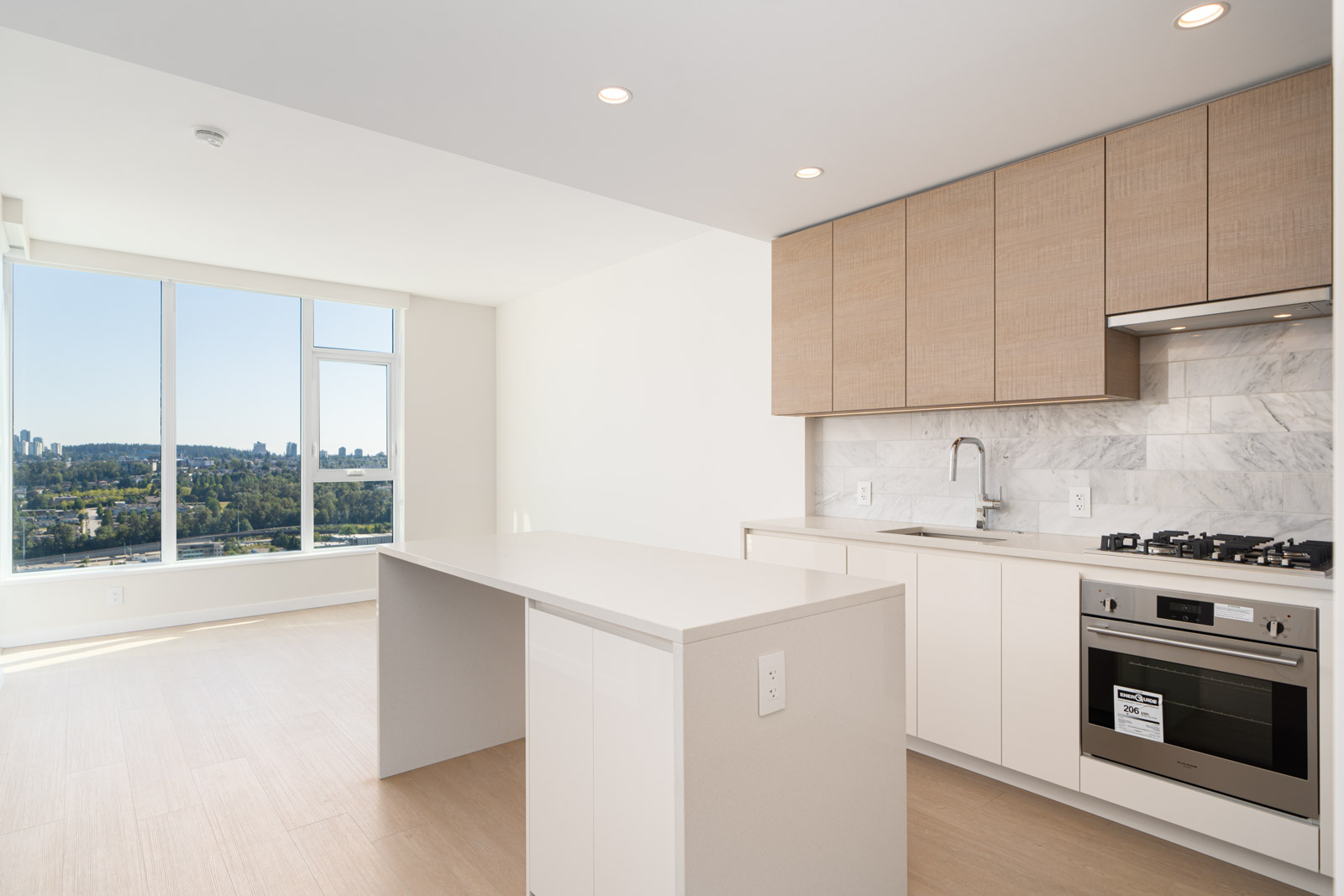 Modern kitchen with a marble backsplash, wood cabinets, and an island. Oven and stovetop are built-in. Large window shows a cityscape view with trees and buildings.