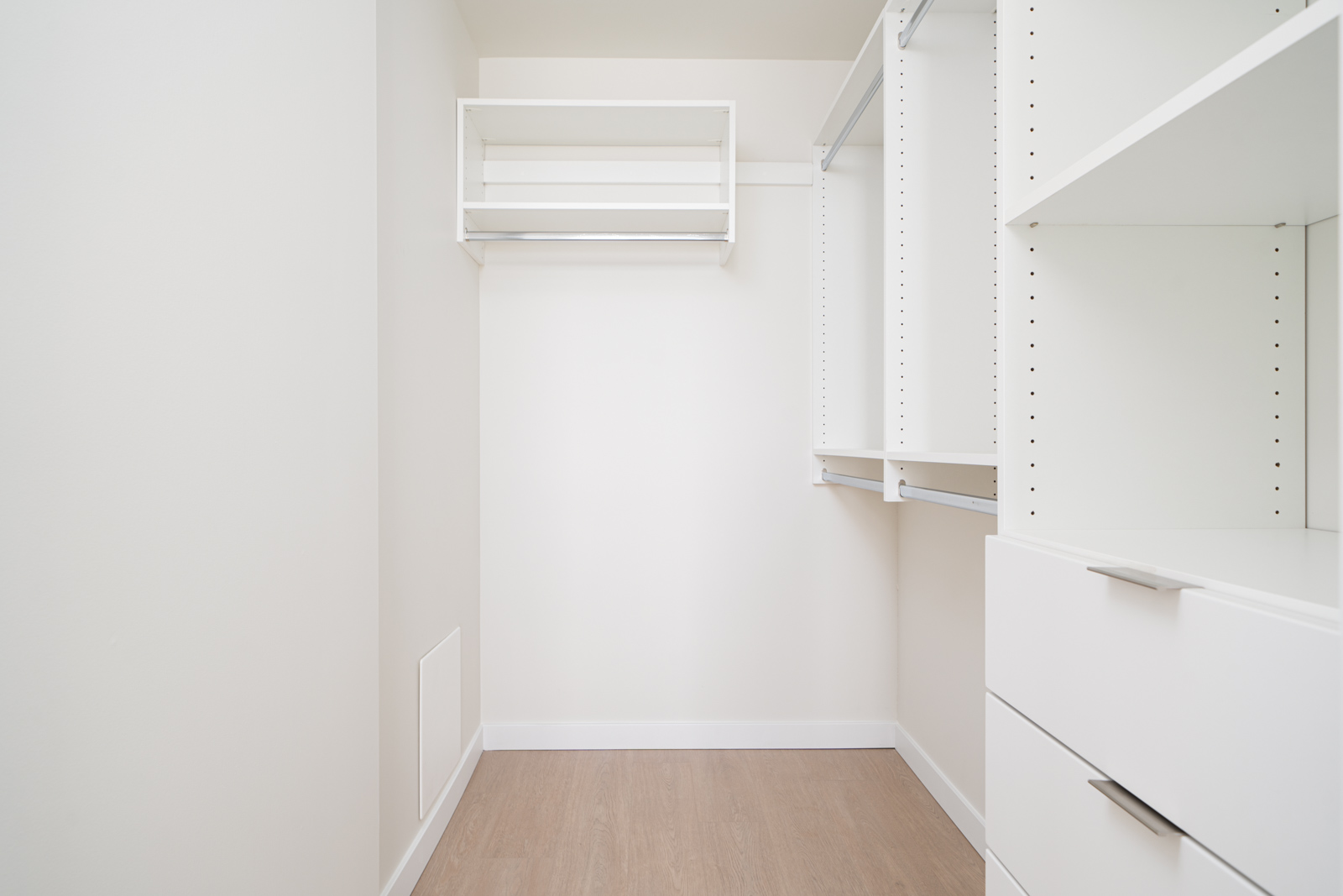 Empty walk-in closet with wooden flooring, white shelving units, and a hanging rod against a light-colored wall.