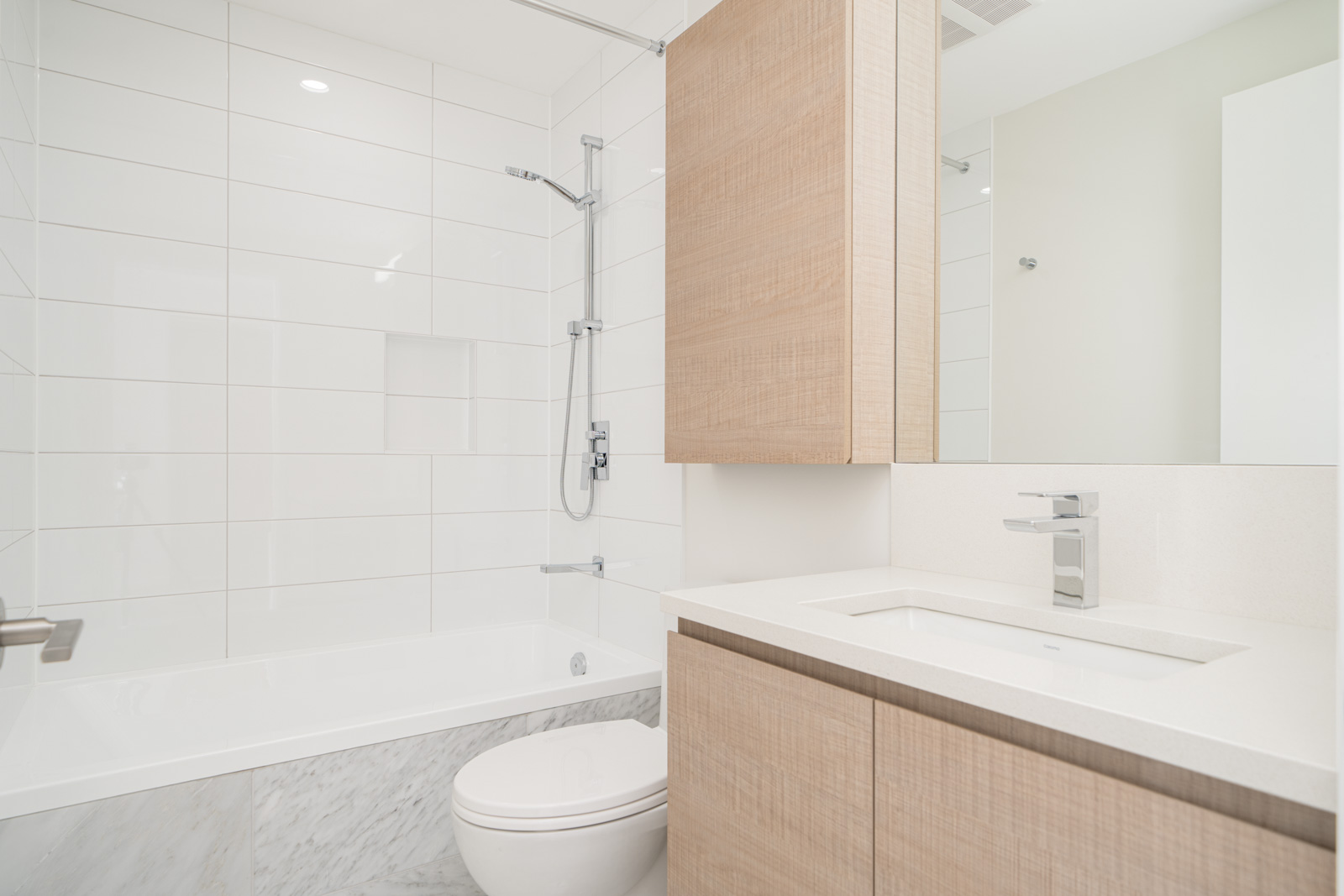 Modern bathroom with a white bathtub, wall-mounted shower, toilet, and wooden cabinet with a sink. White tiled walls and a large mirror are present.
