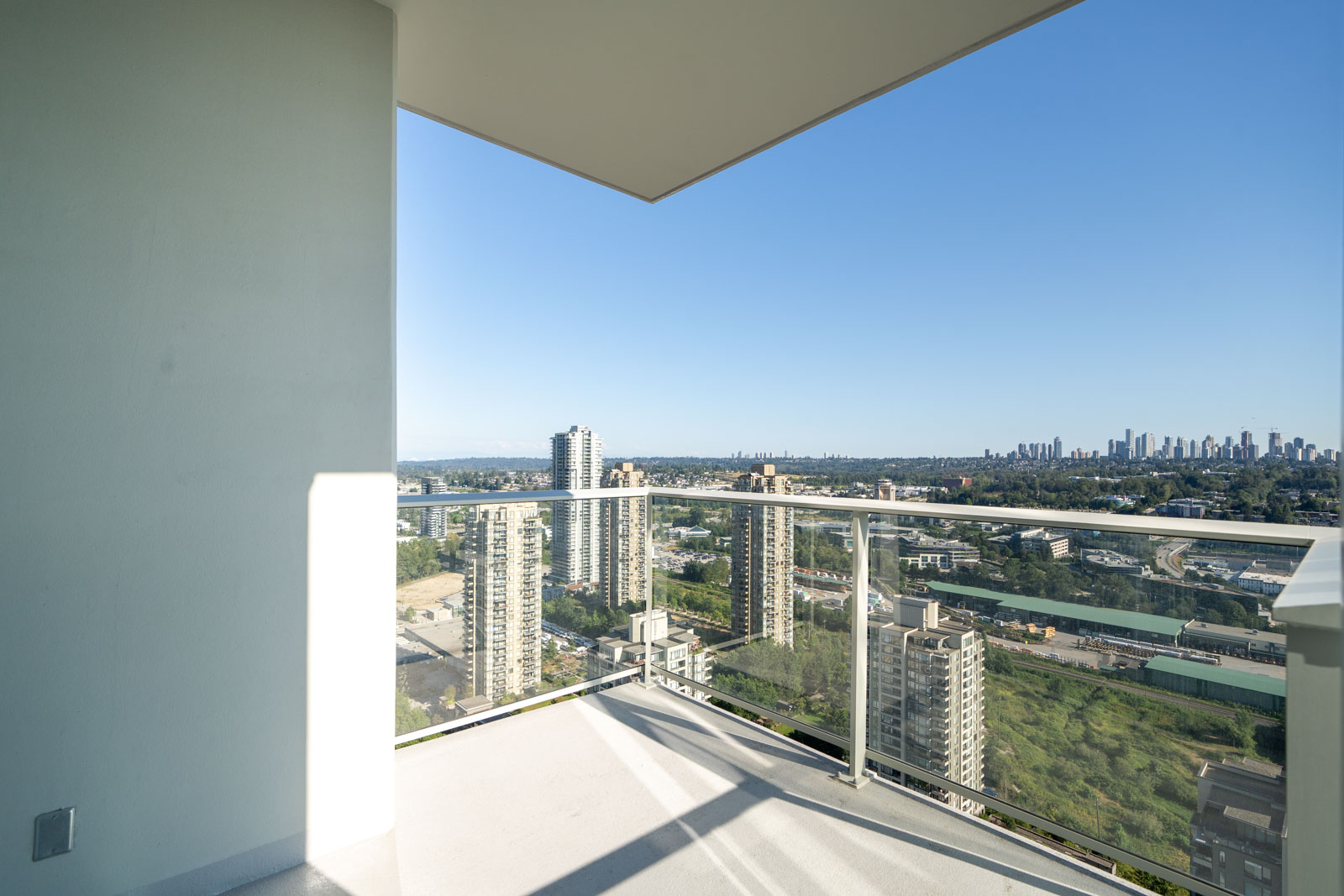 View from a balcony showing a cityscape with multiple high-rise buildings, trees, and a clear blue sky in the background.