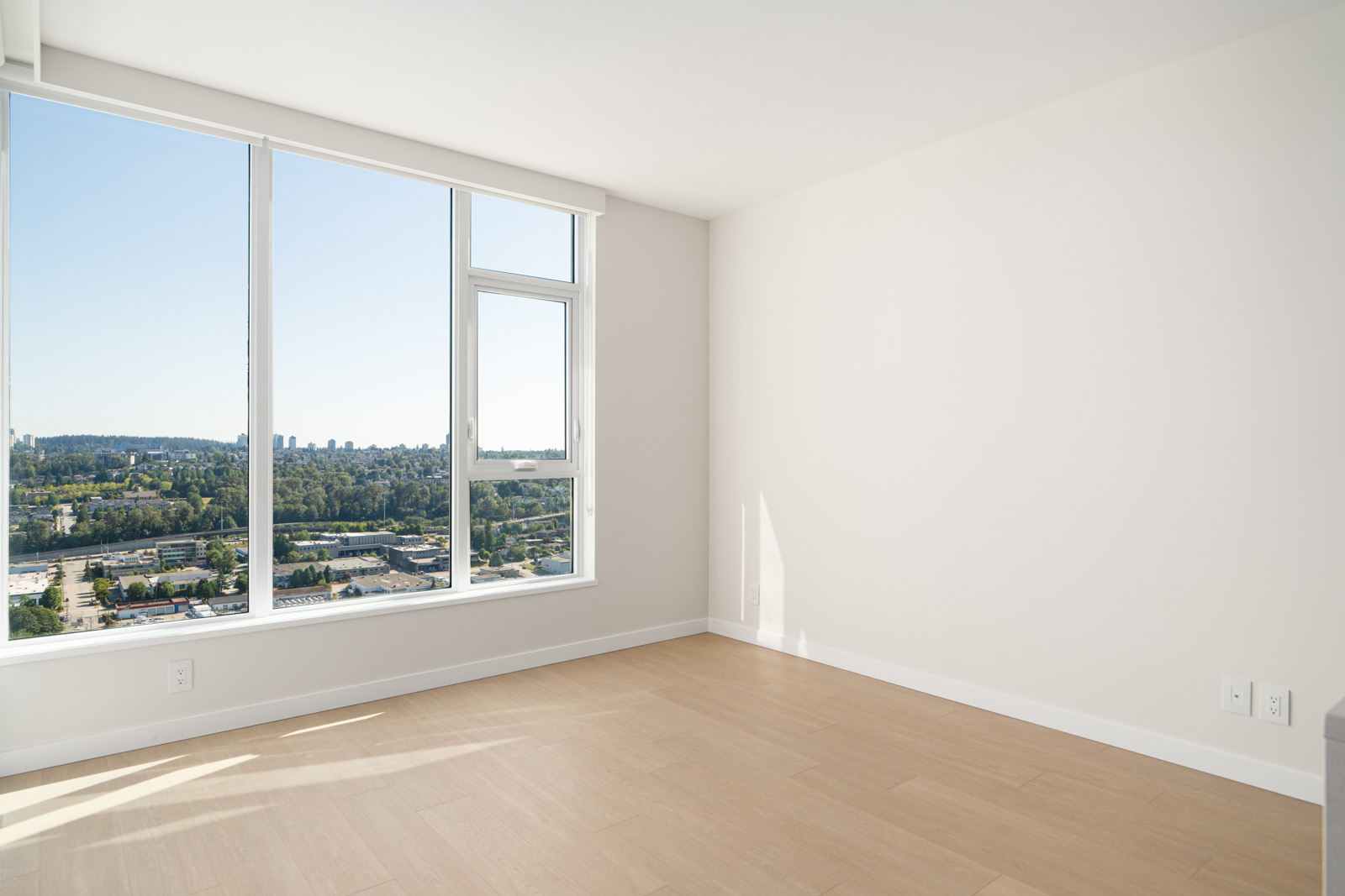Empty room with light wood flooring and large windows offering a view of a city and trees under a clear blue sky. Walls are painted white, and sunlight illuminates the space.