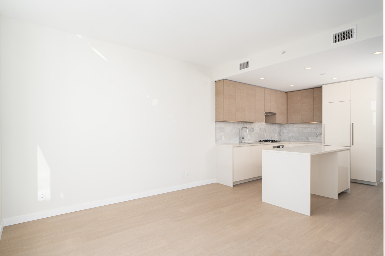 Modern kitchen with light wood flooring, white walls, and natural wood cabinets. Features an island, marble backsplash, built-in stove, and refrigerator. Bright natural lighting from the left.