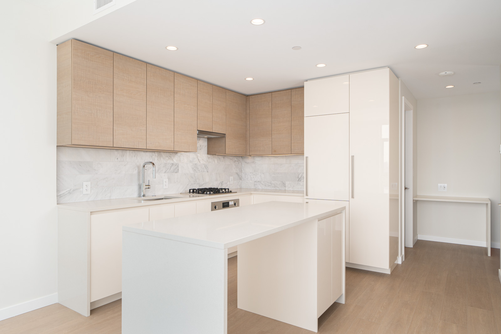 Modern kitchen with light wood cabinets, white countertops, and a central island. Marble backsplash and built-in appliances are visible, with recessed lighting illuminating the space.