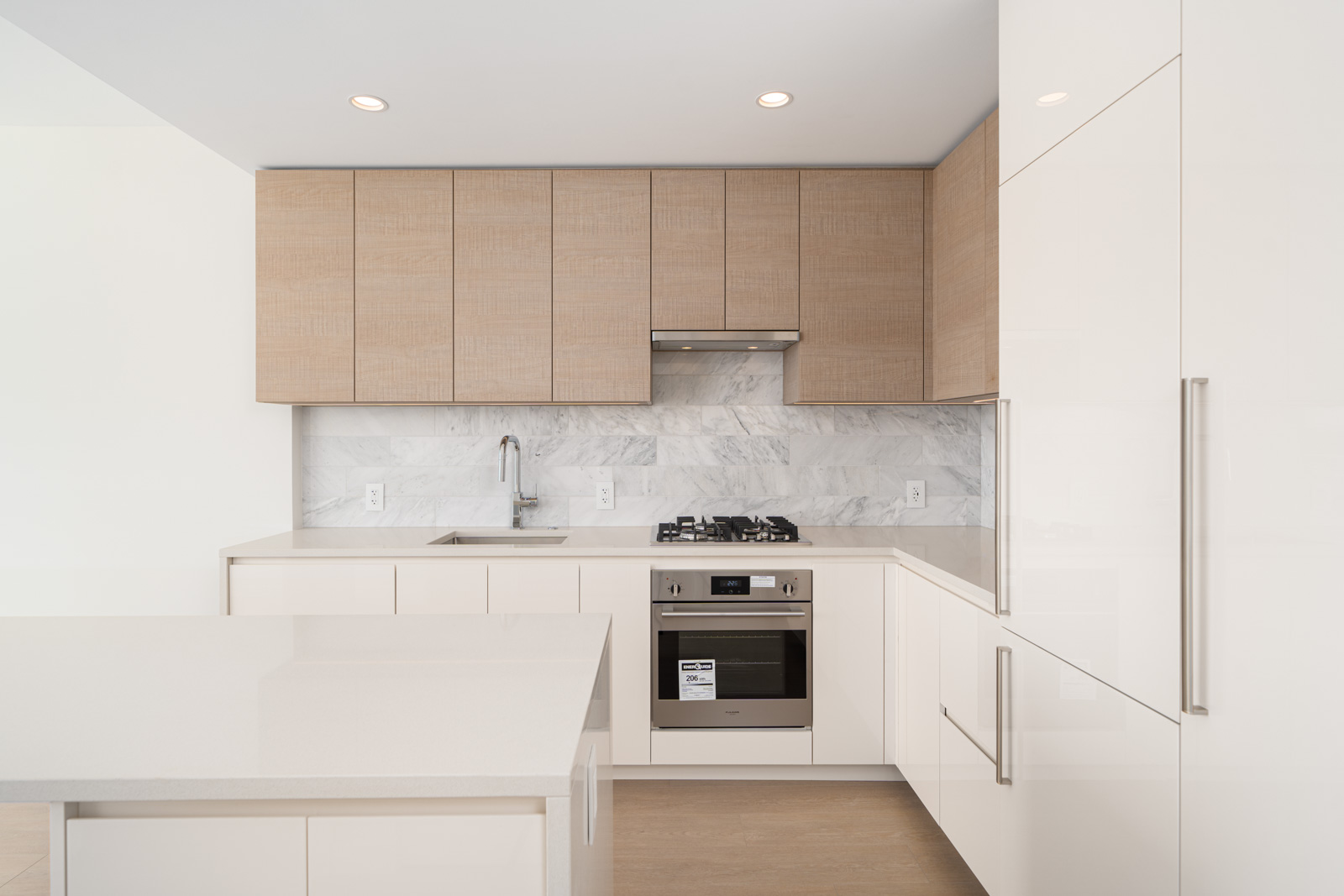 Modern kitchen with light wood cabinets, marble backsplash, stainless steel oven, gas stovetop, and white countertops. Minimalist design with recessed lighting.