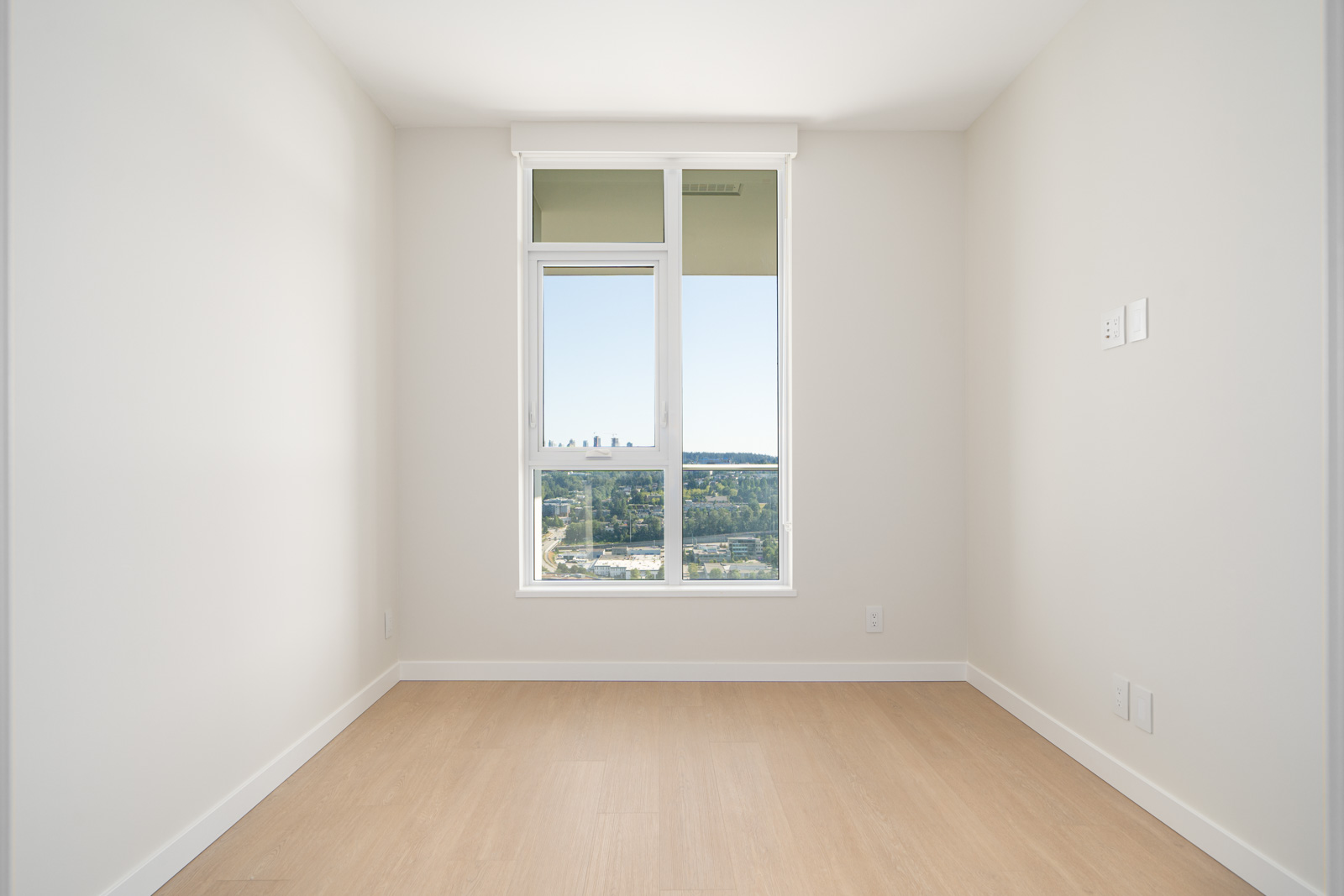 An empty room with beige walls and wooden floor, featuring a large window showing a cityscape and greenery outside.
