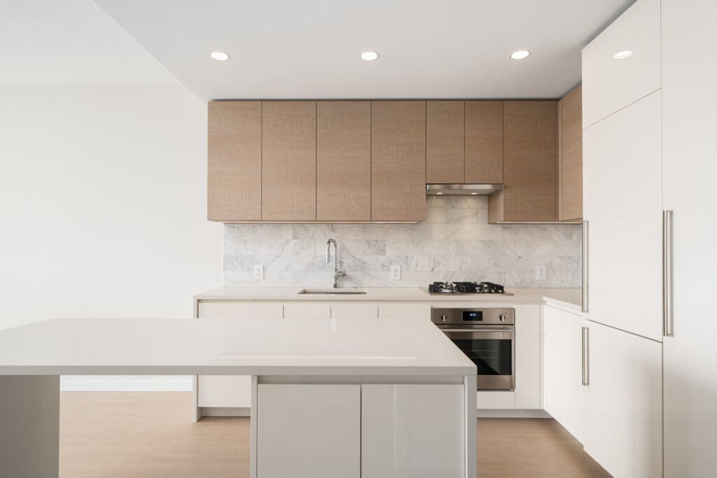 A modern kitchen with light wood cabinets, white countertops, a marble backsplash, a stainless steel stove and hood, built-in oven, sink, and an island.