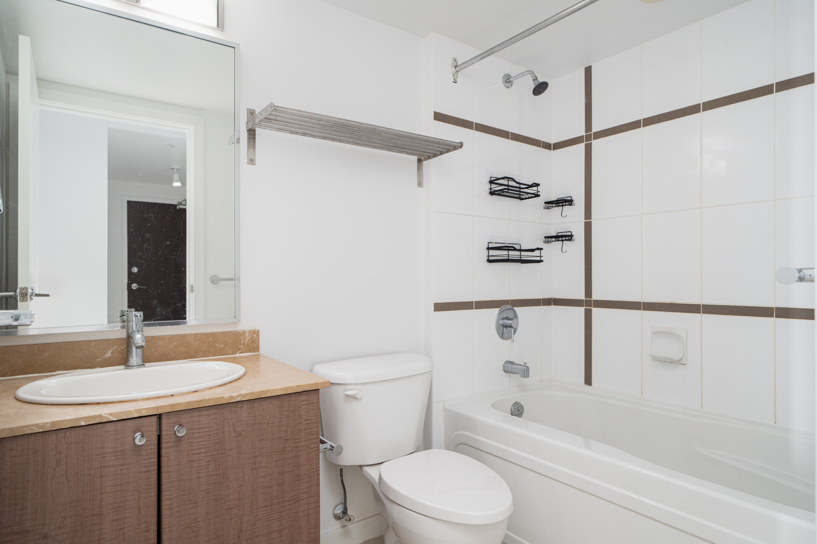 A modern bathroom with a mirror, sink, toilet, and bathtub. The walls are white with brown tile accents, and there are black wire shelves in the bathtub area.