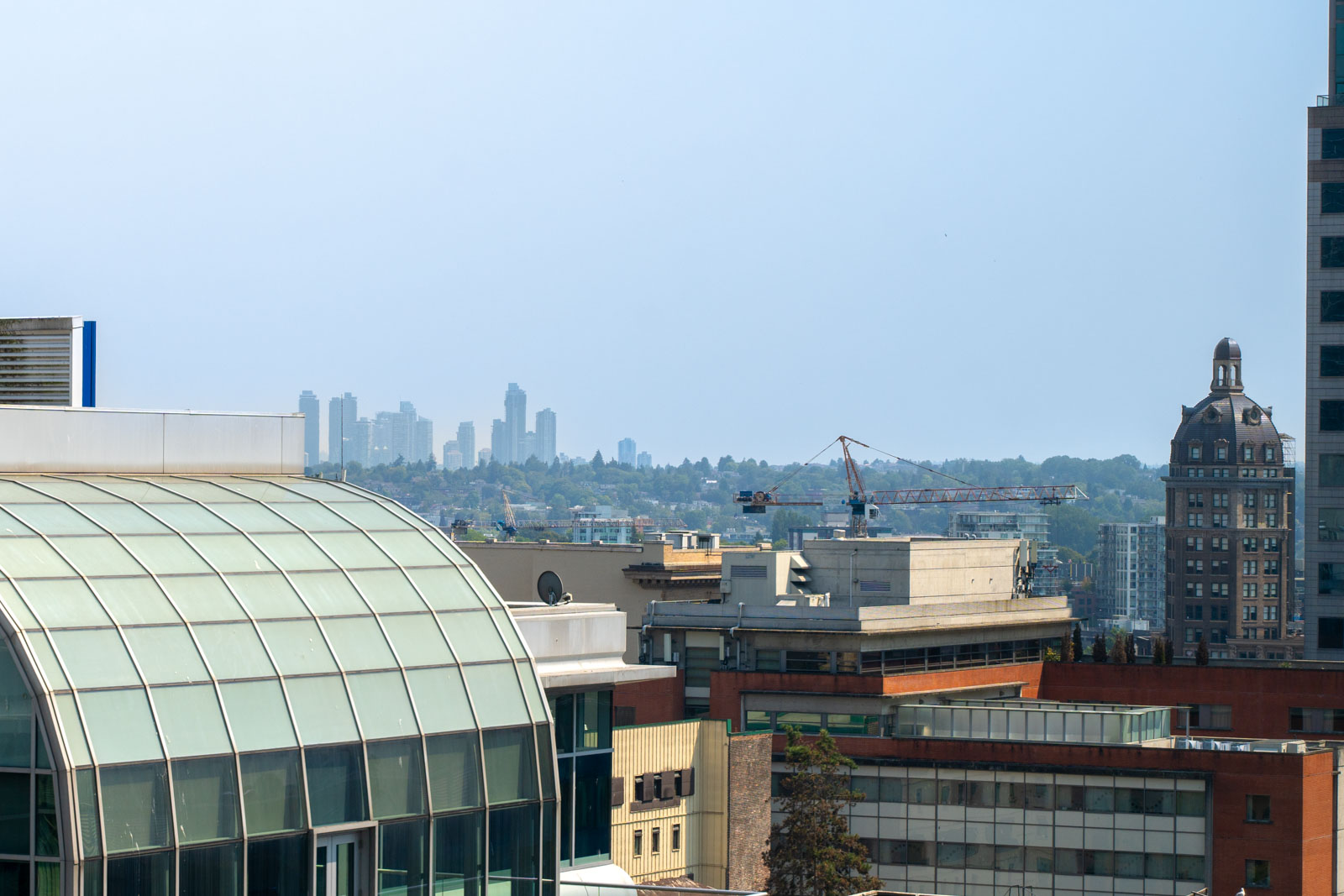 A view from the window looking out at the Vancouver skyline. Modern and older buildings, a glass-roofed structure in the foreground, a construction crane, and high-rise buildings in the distance are visible under a clear blue sky.