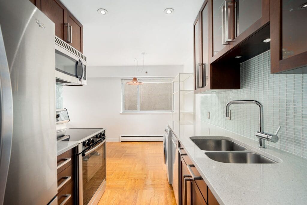 A modern kitchen with stainless steel appliances, under-cabinet lighting, a dual-basin sink, brown cabinetry, light gray countertops, and a window with a light fixture above.