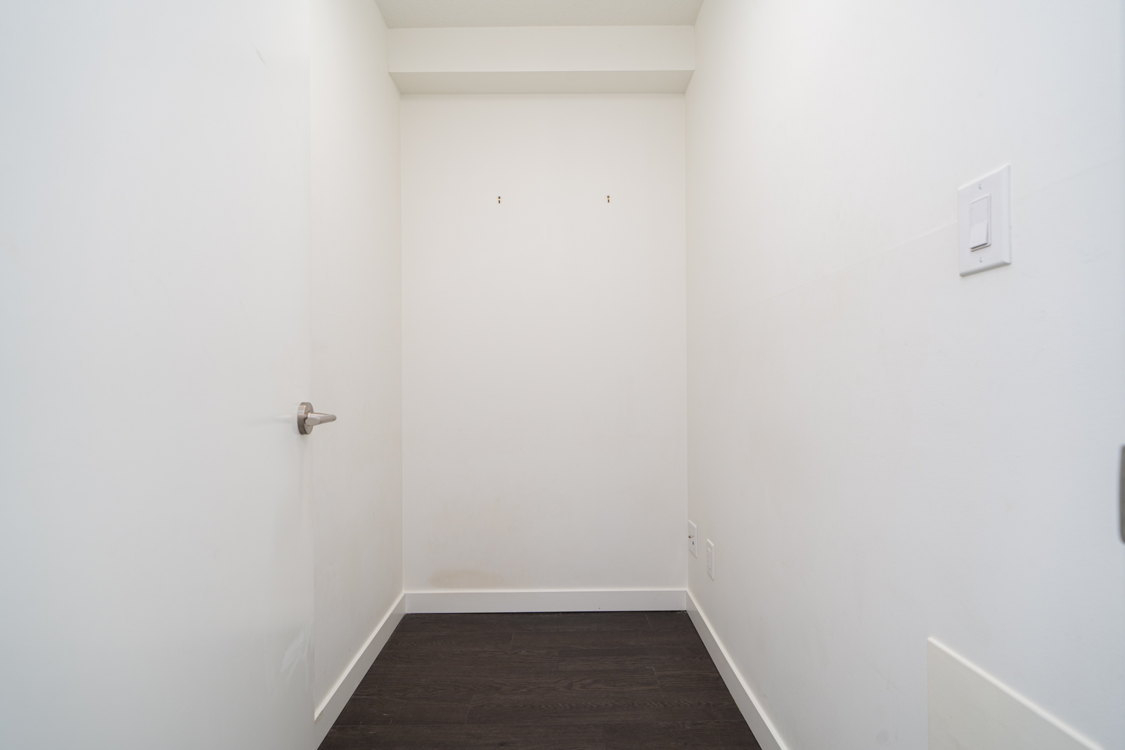 Small empty room with white walls, a white door on the left, dark wood floor, and a light switch on the right wall.