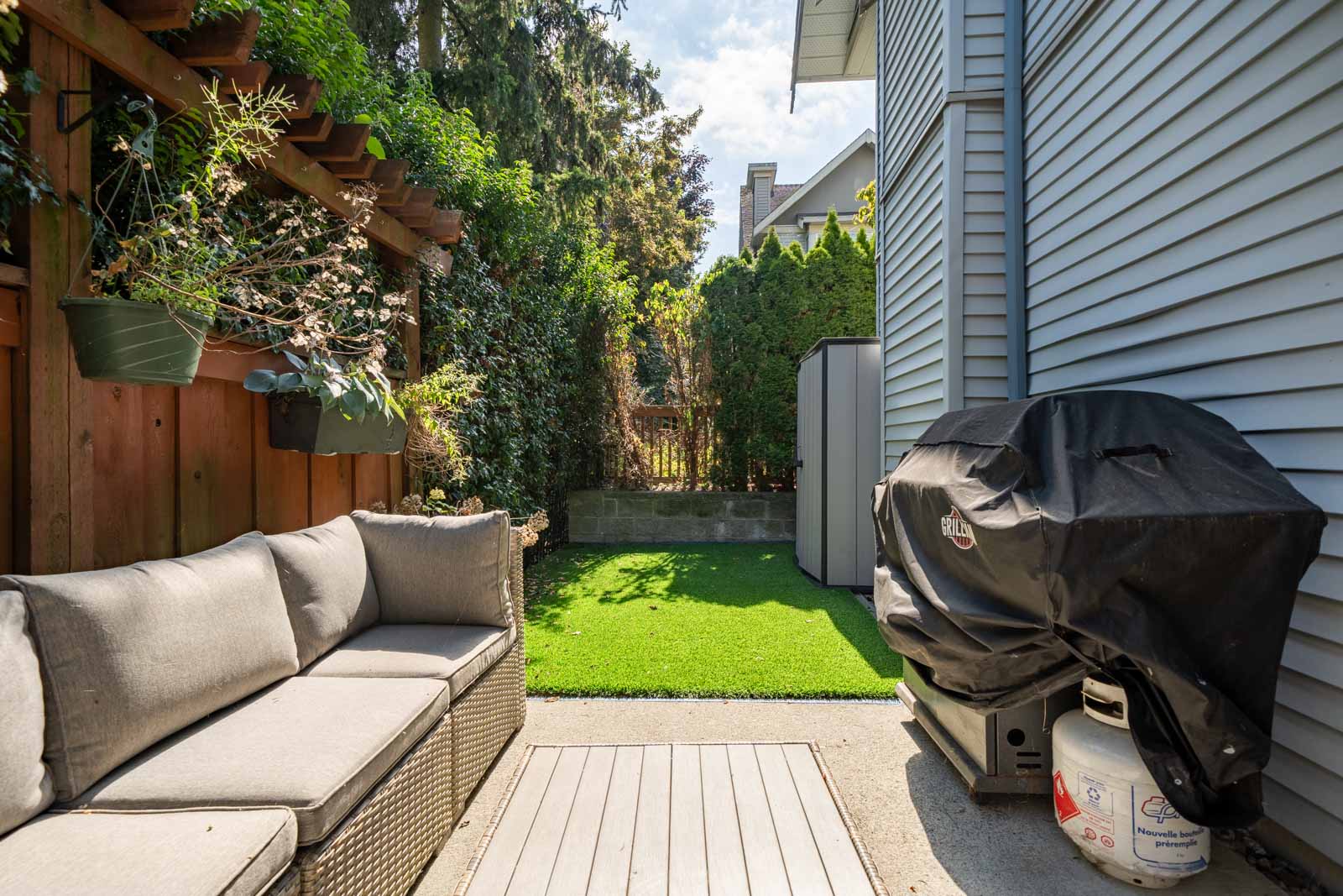 A backyard with patio furniture, a grill, and a green lawn. There is a wooden fence with plants on the left and a building with siding on the right.