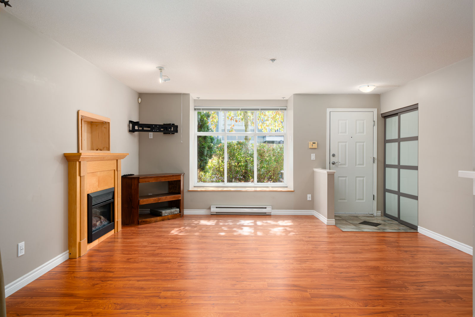 A living room with hardwood floors, a fireplace with a wooden mantle, an empty wall-mounted bracket, a wooden shelf, a large window, and an entry door and closet to the right.