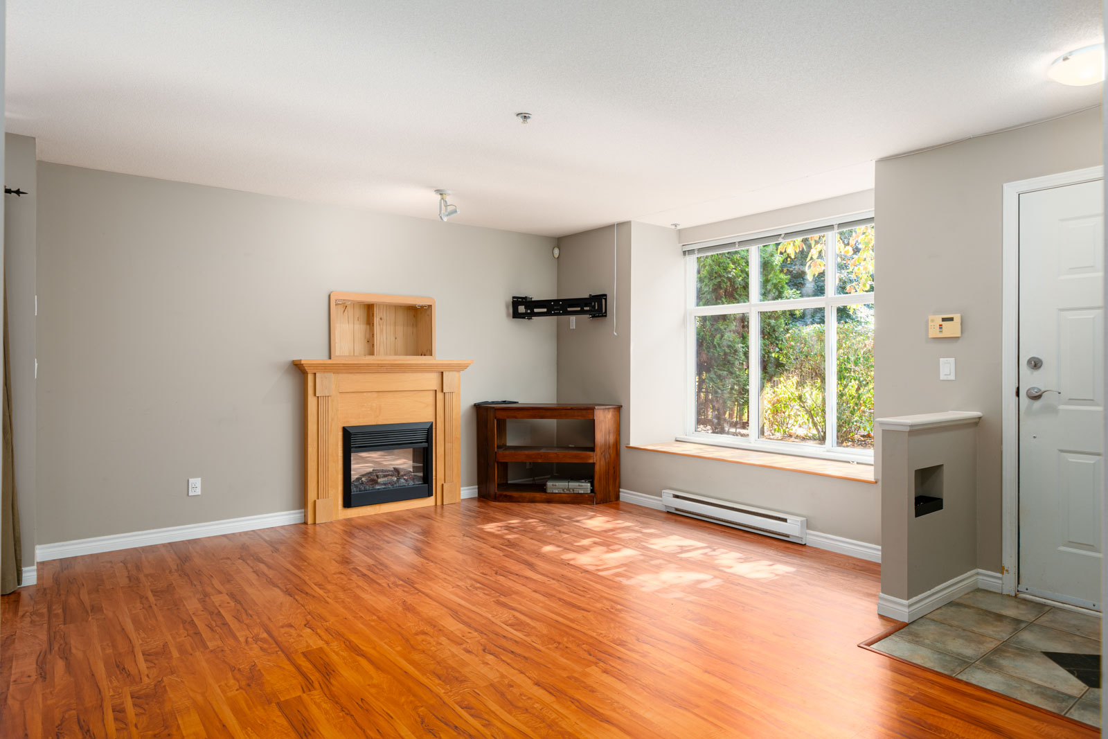 A living room with hardwood floors, a fireplace with a wooden mantle, an empty wall-mounted bracket, a wooden shelf, a large window, and an entry door to the right.