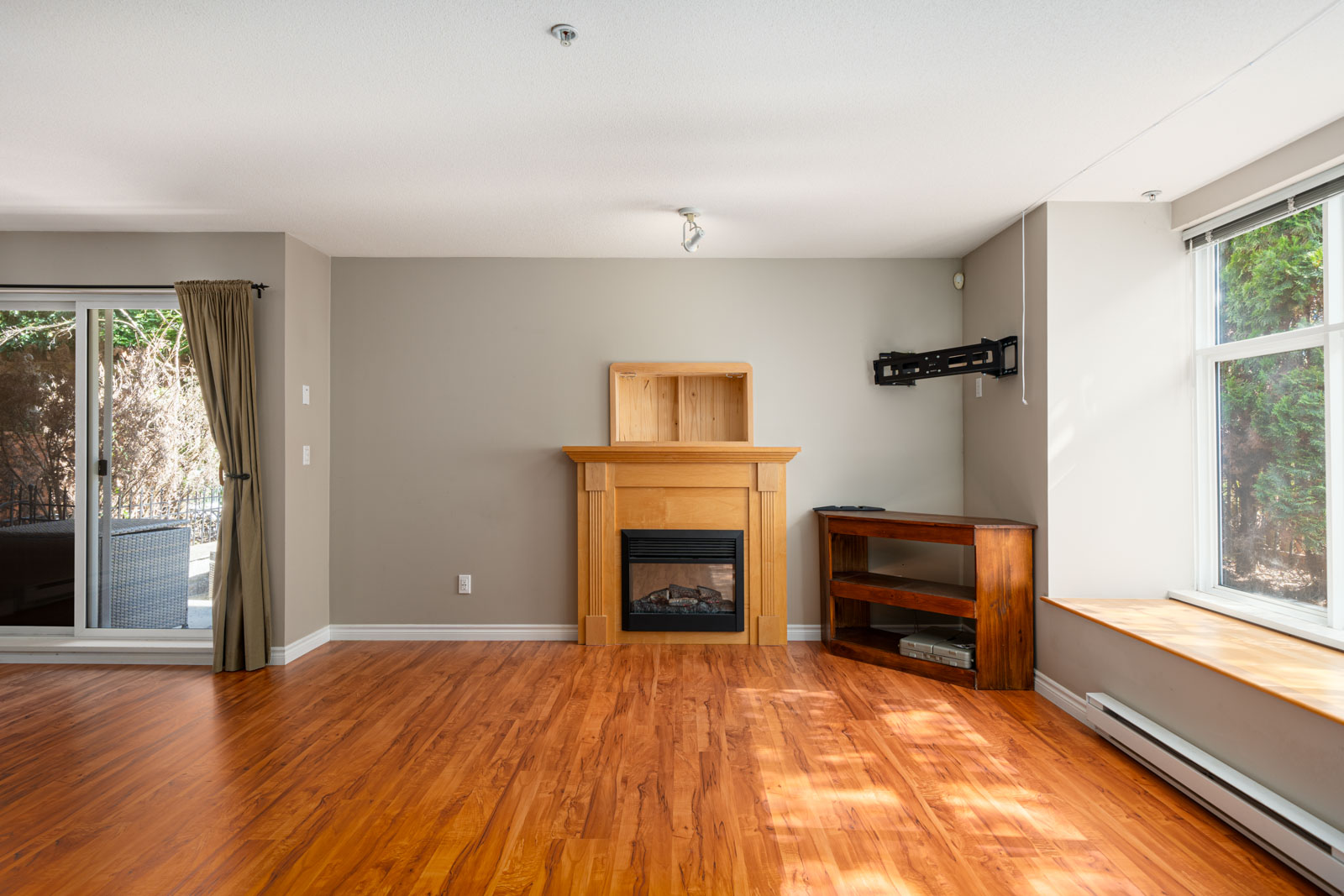 A living room with hardwood floors, a fireplace with a wooden mantle, an empty wall-mounted bracket, a wooden shelf, a large window to the right, and a sliding door with a curtain leading outside to the left.