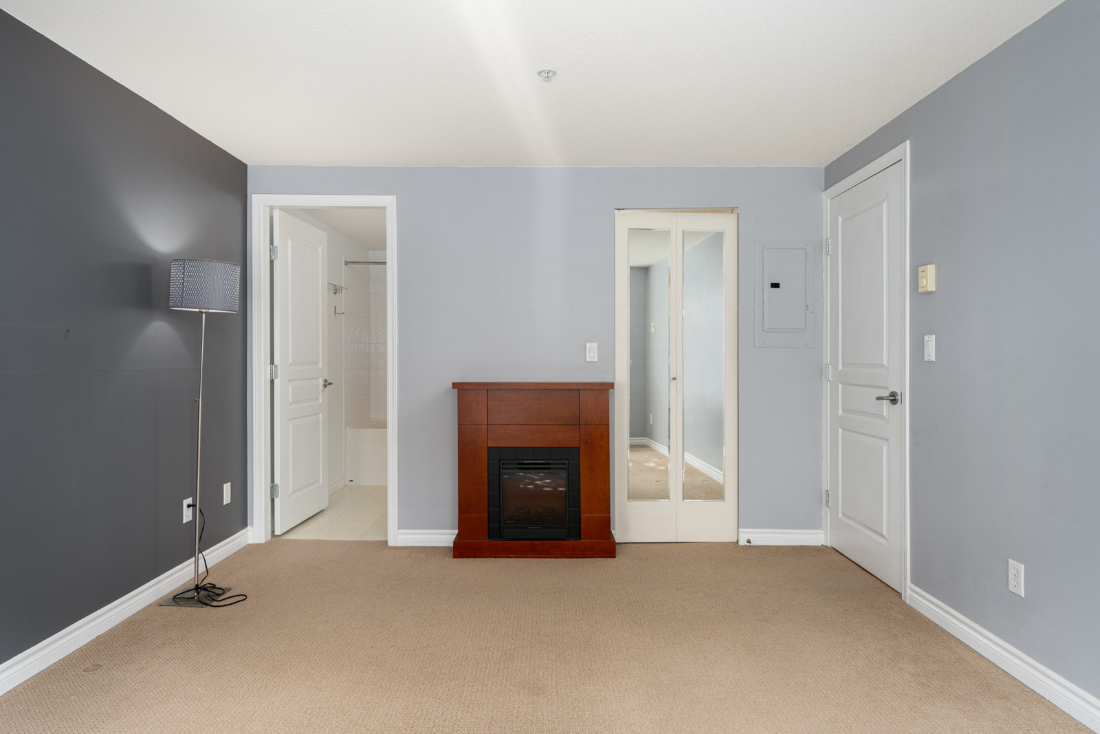 A bedroom with gray walls, beige carpet, a brown electric fireplace, a floor lamp, and two white doors. One door leads to a bathroom, and the other door has a mirrored surface.