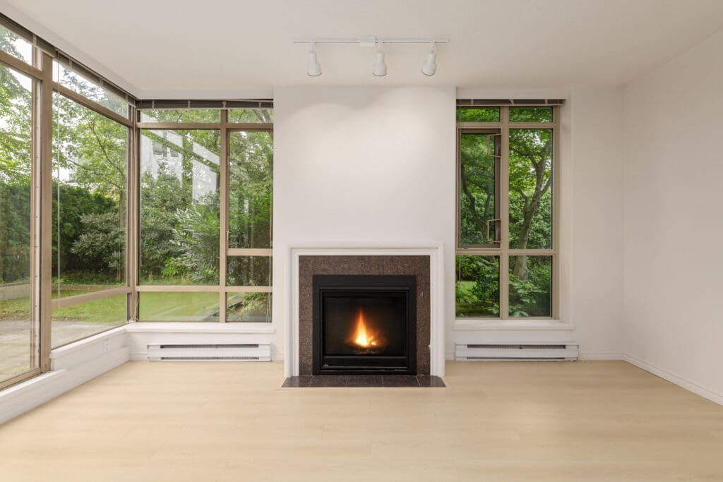 A modern living room with large windows, a white wall, and a central fireplace. The room has light-colored wooden flooring and outdoor greenery is visible through the windows.
