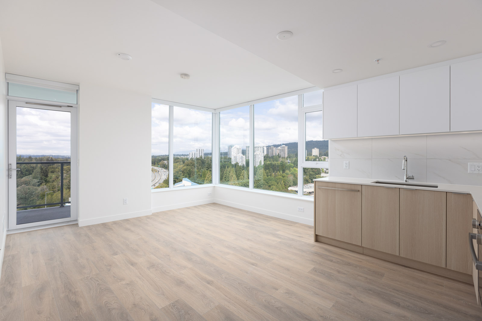 A modern, empty apartment with light hardwood floors, large windows offering a scenic view, and a kitchenette with white and wood-toned cabinets.