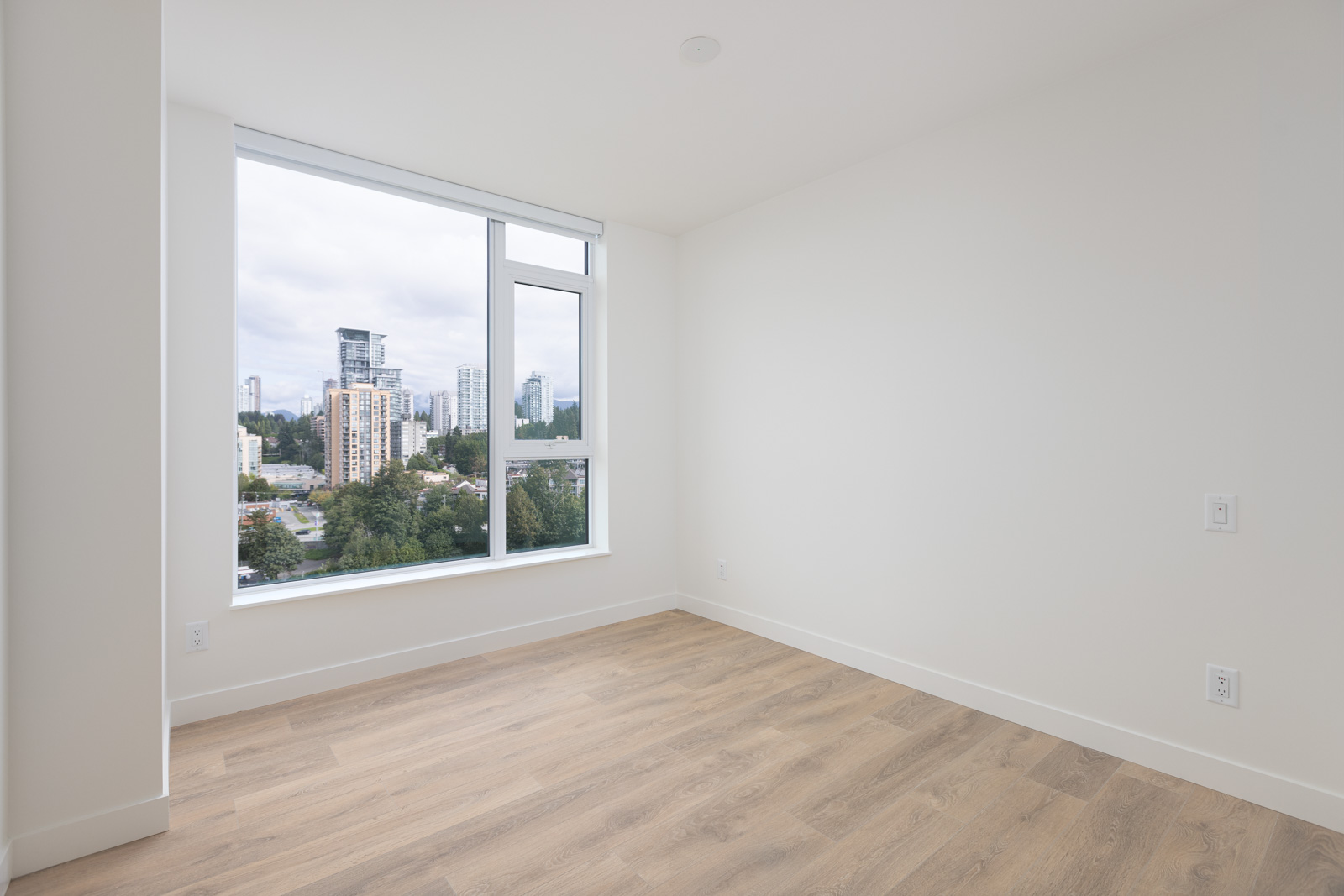 An empty room with light wood flooring, a large window displaying a cityscape with tall buildings and trees outside. White walls and a ceiling light.