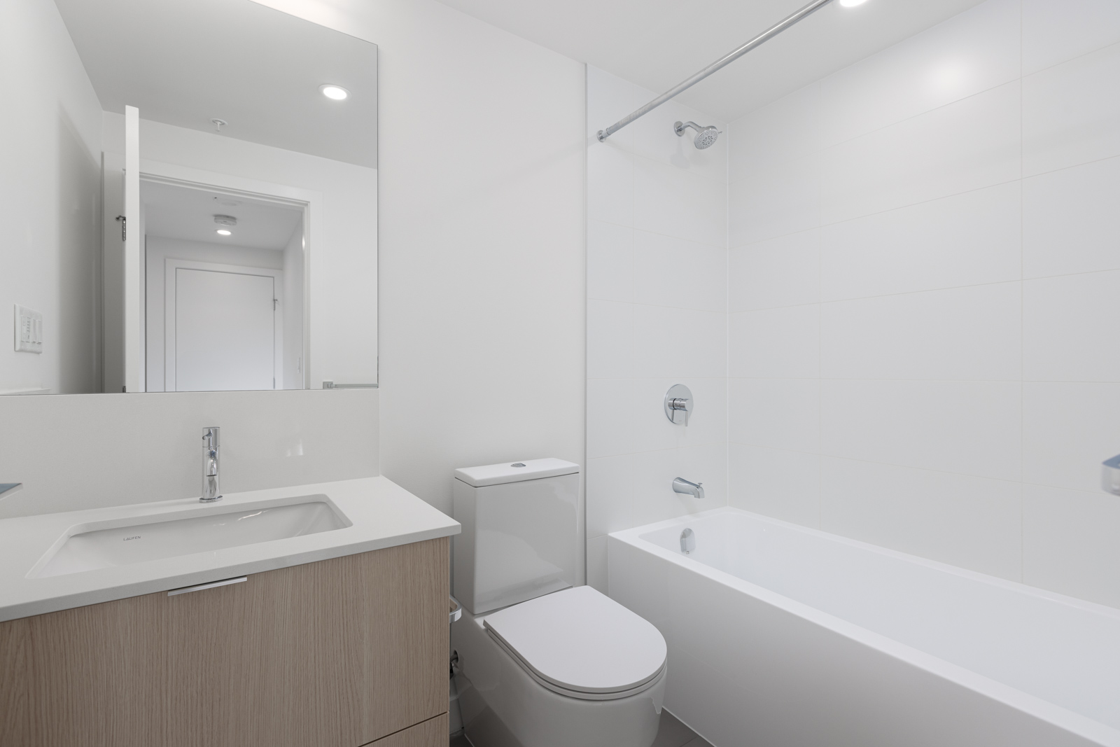 Modern bathroom with white walls, a bathtub with an overhead shower, a toilet, and a wooden vanity with a sink and mirror.