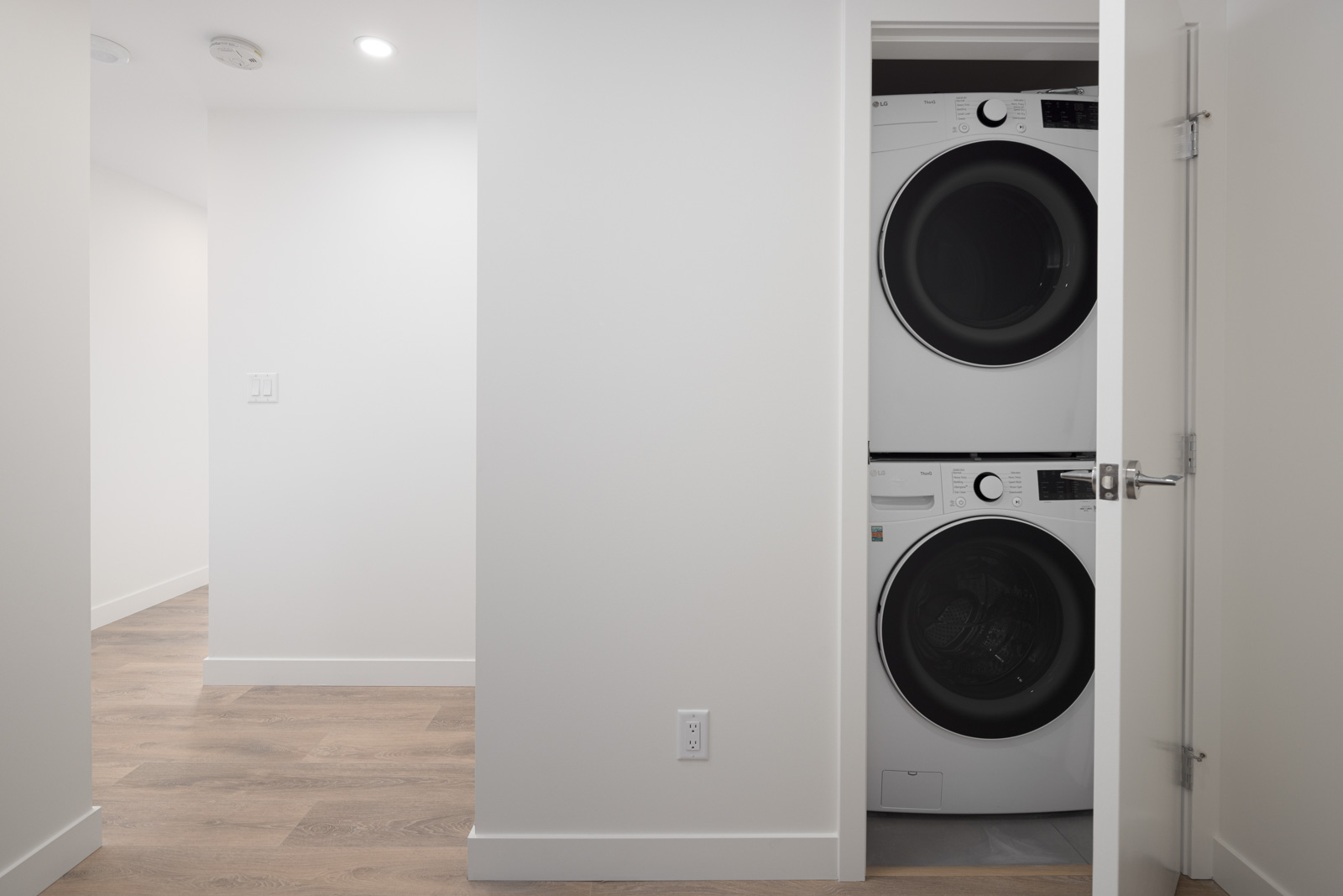 A small laundry closet with a stacked washer and dryer, partially open door, located in a minimalist, bright white room with wooden floors.