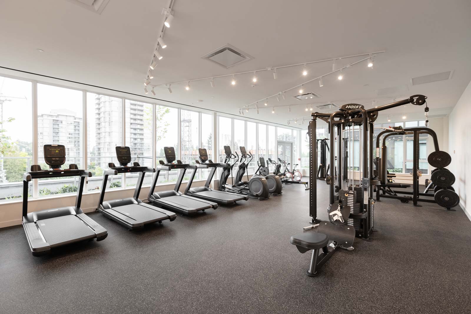 A modern gym with treadmills, exercise bikes, weight machines, and free weights, with large windows and a view of a cityscape.