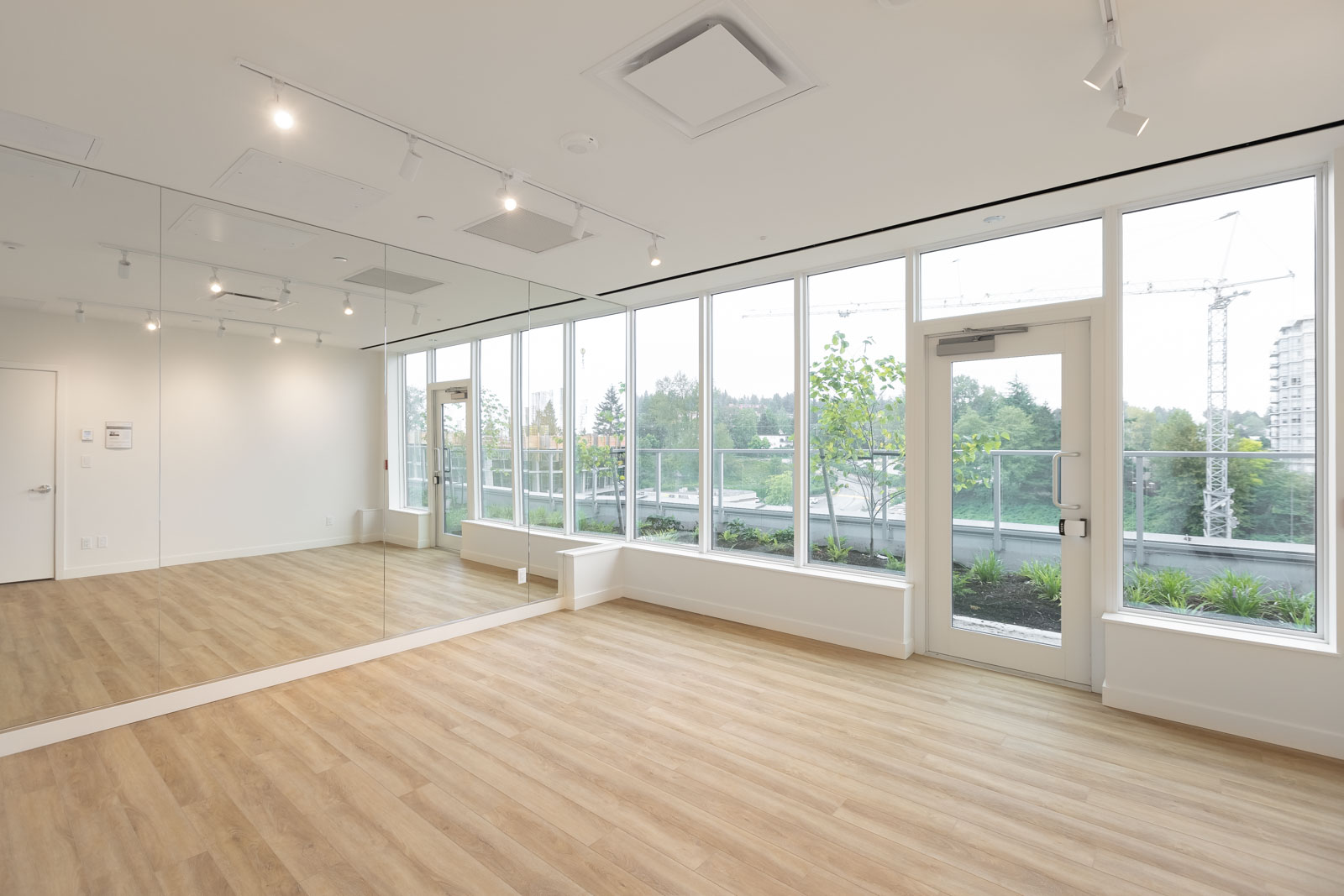 A spacious room with wooden flooring, large mirrors on one wall, and floor-to-ceiling windows offering a view of trees and cranes in the distance. Track lighting is installed on the ceiling.