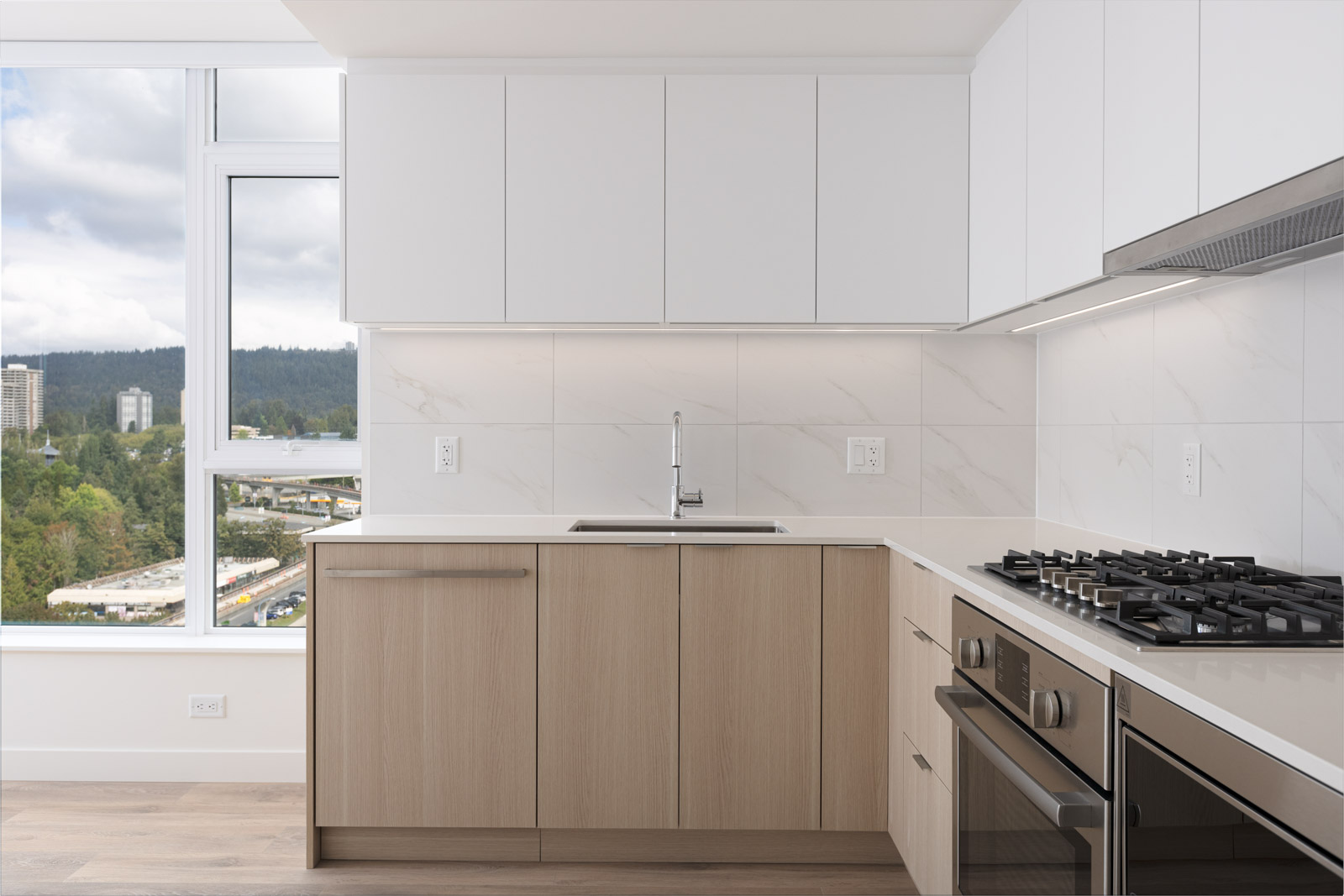 Modern kitchen with light wood lower cabinets, white upper cabinets, and stainless steel appliances. Large window offers a scenic view of trees and buildings outside.