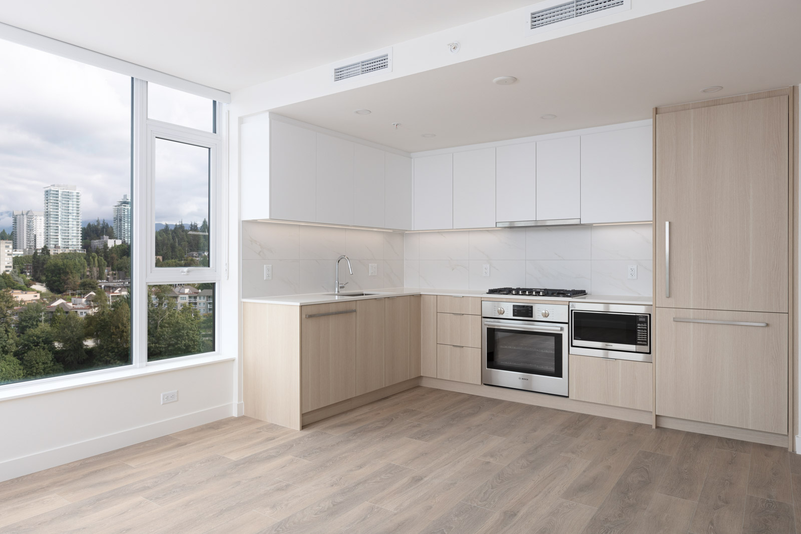 Modern kitchen with light wood cabinetry, white countertops, stainless steel appliances, and large windows offering a city and tree-lined view.