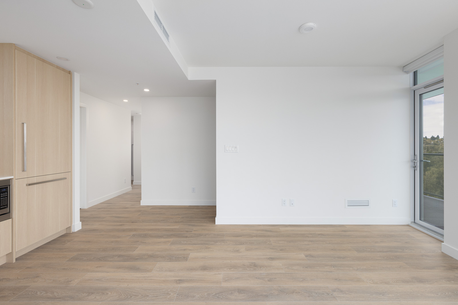 A modern, unfurnished apartment with white walls, wood flooring, a built-in kitchen on the left, a doorway leading to another room, and a large window with a sliding glass door on the right.